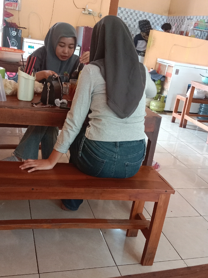 Two women are seated at a wooden table and bench in what appears to be a casual dining space. Both women are wearing hijabs. One faces the camera, while the other has her back to the viewer. Various items are on the table, including drinks, containers of utensils, and a small purse. Other individuals are visible in the background. The room has a casual ambiance with white tiled floors and basic furnishings.