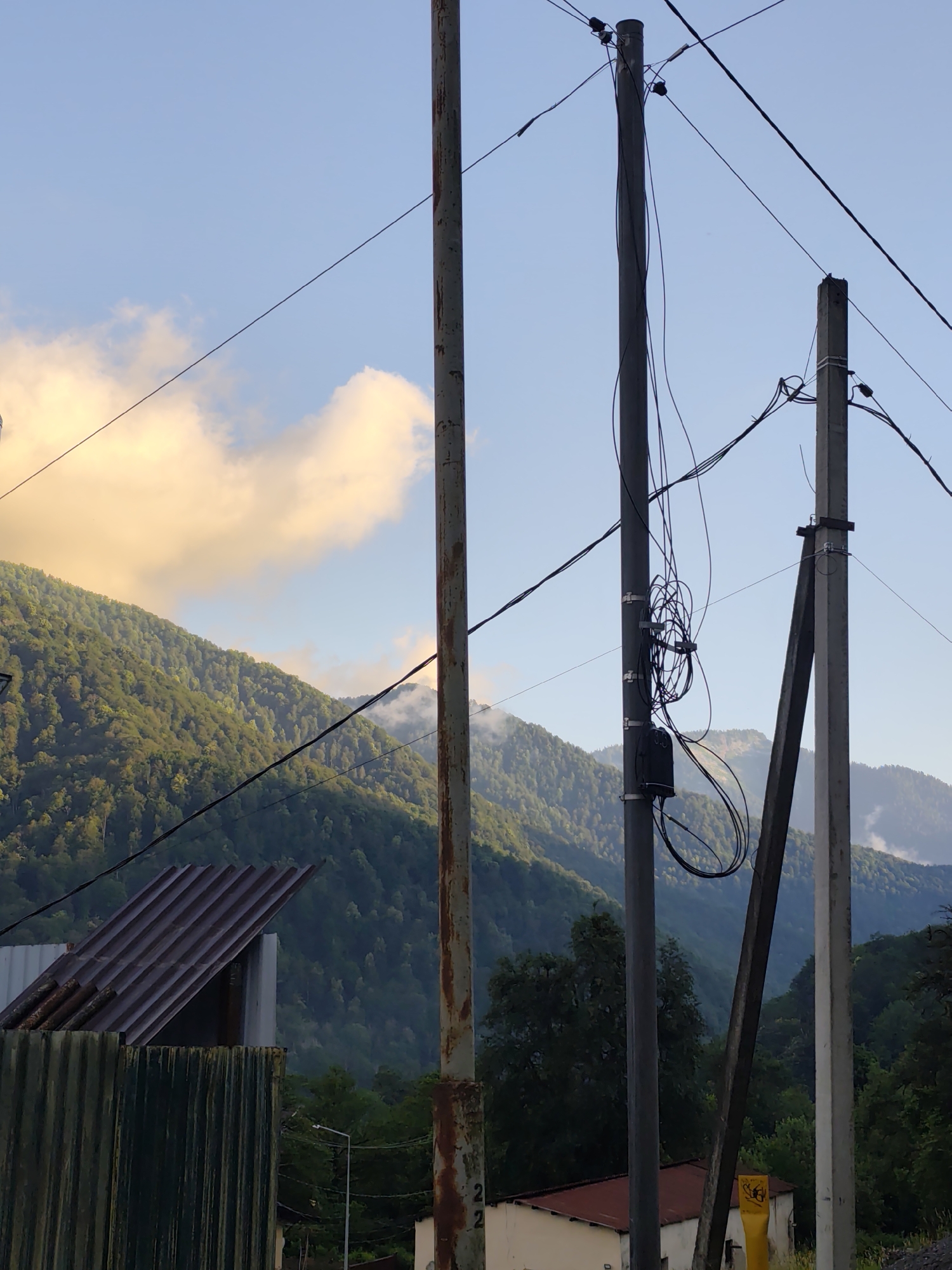 A view of a mountainous landscape in Krasnaya Polyana, Russia, is seen under a clear blue sky with some scattered, golden-tinged clouds. In the foreground, several utility poles with a tangle of wires and a black box are prominent. To the left, part of a corrugated metal structure and a brown, corrugated metal roof are visible. The background is dominated by lush green, forested mountains, with some areas shrouded in mist. The overall scene depicts a rural or semi-rural setting with infrastructure for electricity or communication. There are no people visible in the image.