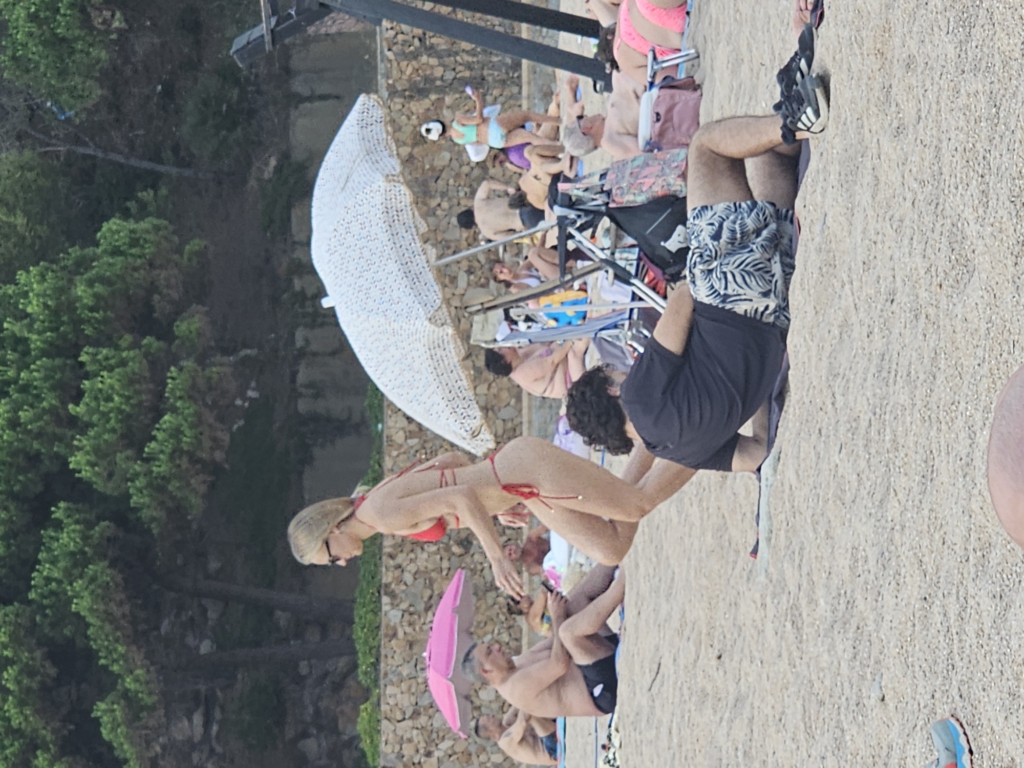 This image captures a beach scene in Blanes, Spain, with several individuals enjoying a sunny day. In the foreground, a person wearing a black t-shirt and patterned shorts is lying on the sand, with athletic shoes nearby. Behind them, a woman in a red bikini is walking, with a large white umbrella to her right. Further back, other beachgoers are visible, some sitting under pink and white umbrellas, and others relaxing on towels or chairs. The environment is sandy, with trees and a stone wall forming the backdrop, suggesting a coastal setting.
