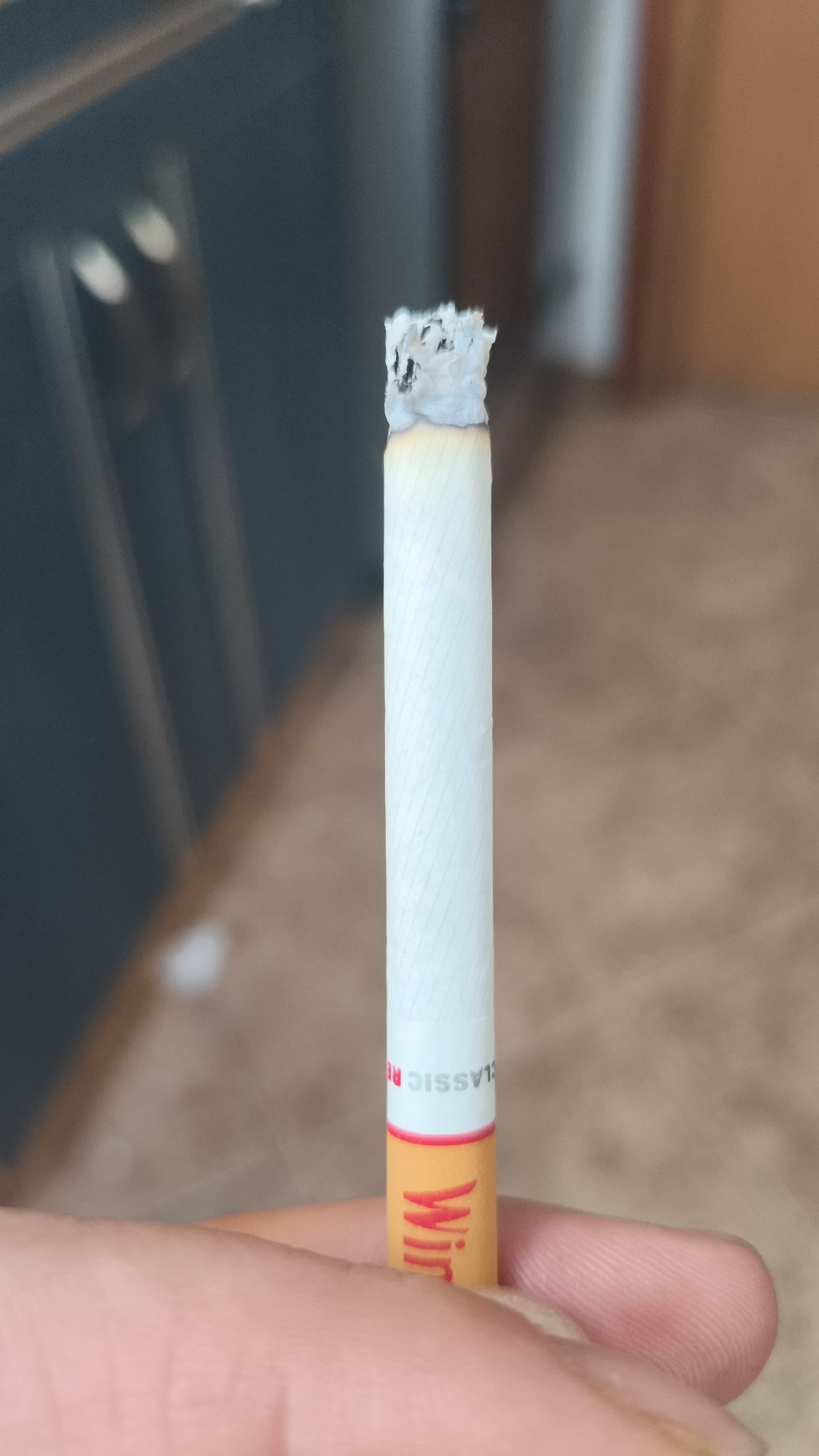 The image shows a burning cigarette being held between a person's fingers. The brand appears to be "Winston Classic R." The background is blurry, making it difficult to discern the exact location, but it seems to be an indoor environment. There's no indication of this image being taken in Nowy Tomyśl, Poland.