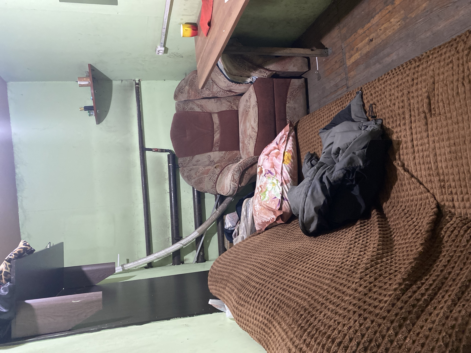 The image shows a sparsely furnished room, likely a small apartment or dorm room, in Novokuznetsk, Russia. The room is dimly lit and appears somewhat cluttered. On the left, a brown textured blanket covers a sofa or bed. A dark gray jacket is draped over it. To the right of the sofa, a large armchair with a floral pattern of brown and pink is visible. In front of the armchair, there is a pink pillow with a floral print. A small wooden table with a checkered cloth is positioned next to the armchair, with a yellow and red mug and a red cloth on it. The walls are painted a pale green. There are pipes running along the wall and a small shelf with some items on it in the corner. The floor is made of wooden planks. The overall impression is of a utilitarian living space.