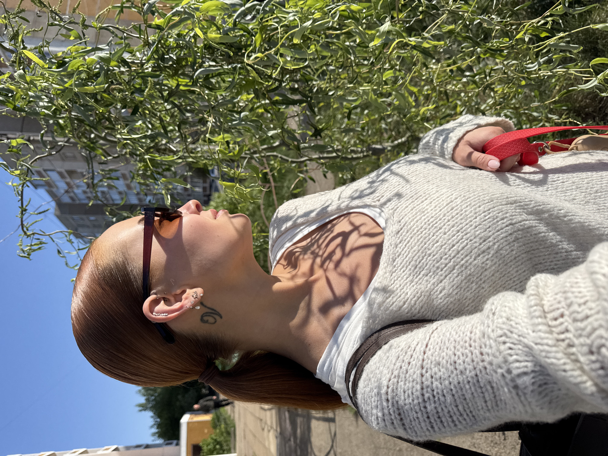 A light-skinned person with reddish-brown hair pulled back into a ponytail stands outdoors in Chaikovsky, Russia. They are wearing a white sweater, a white undershirt, and brown sunglasses, looking up and to the left. A tattoo is visible on their neck, and multiple piercings are on their ear. A red leash is held in their hand. Green foliage and a multistory building are visible in the background. The sky is blue.