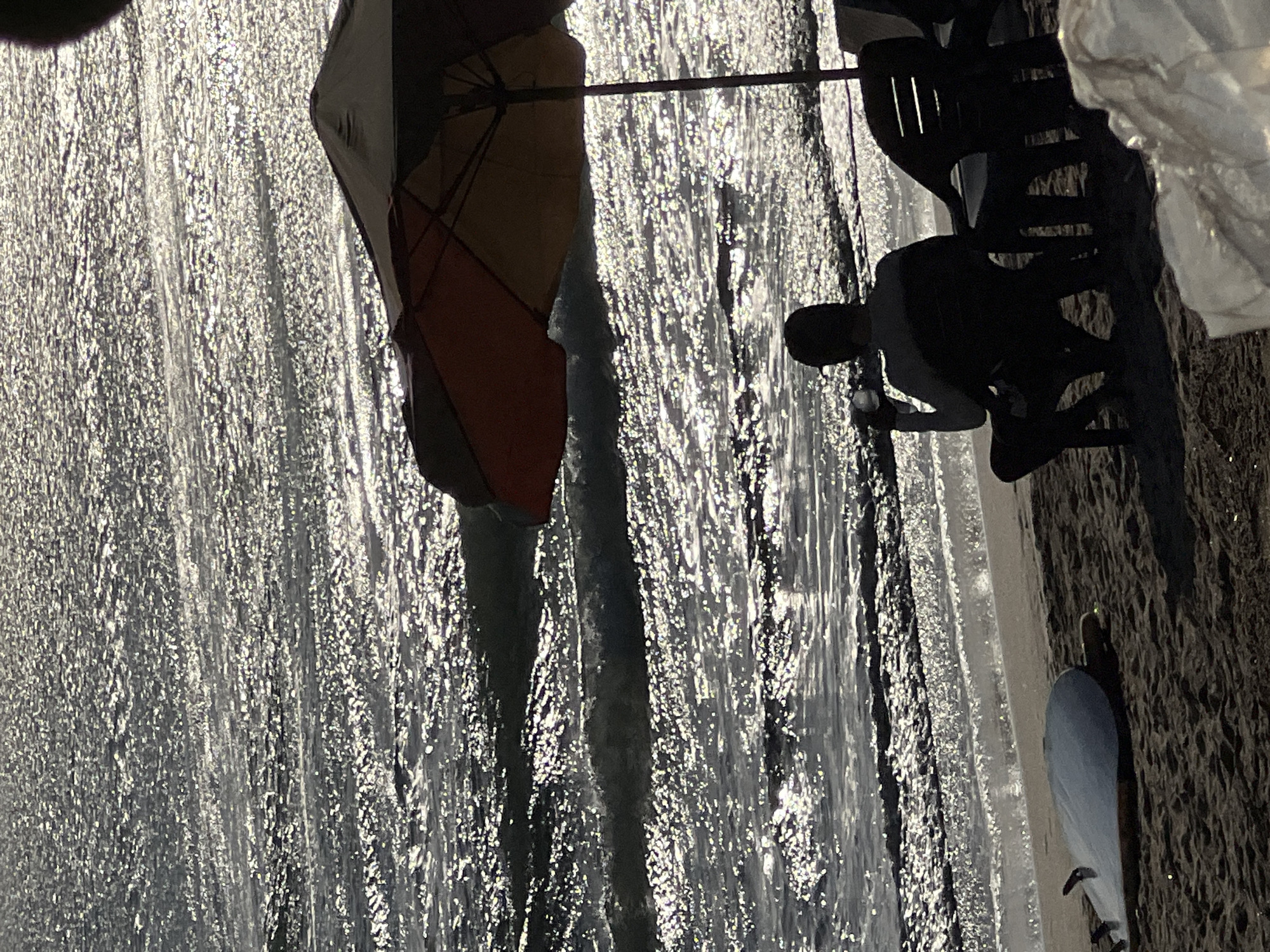 A person is sitting on a beach chair, facing the ocean, holding a drink. The sun is setting, casting a shimmering reflection on the water. A surfboard is lying on the sand near the person. An umbrella is positioned over the chair, providing shade. The scene is set in Kuta, Indonesia.