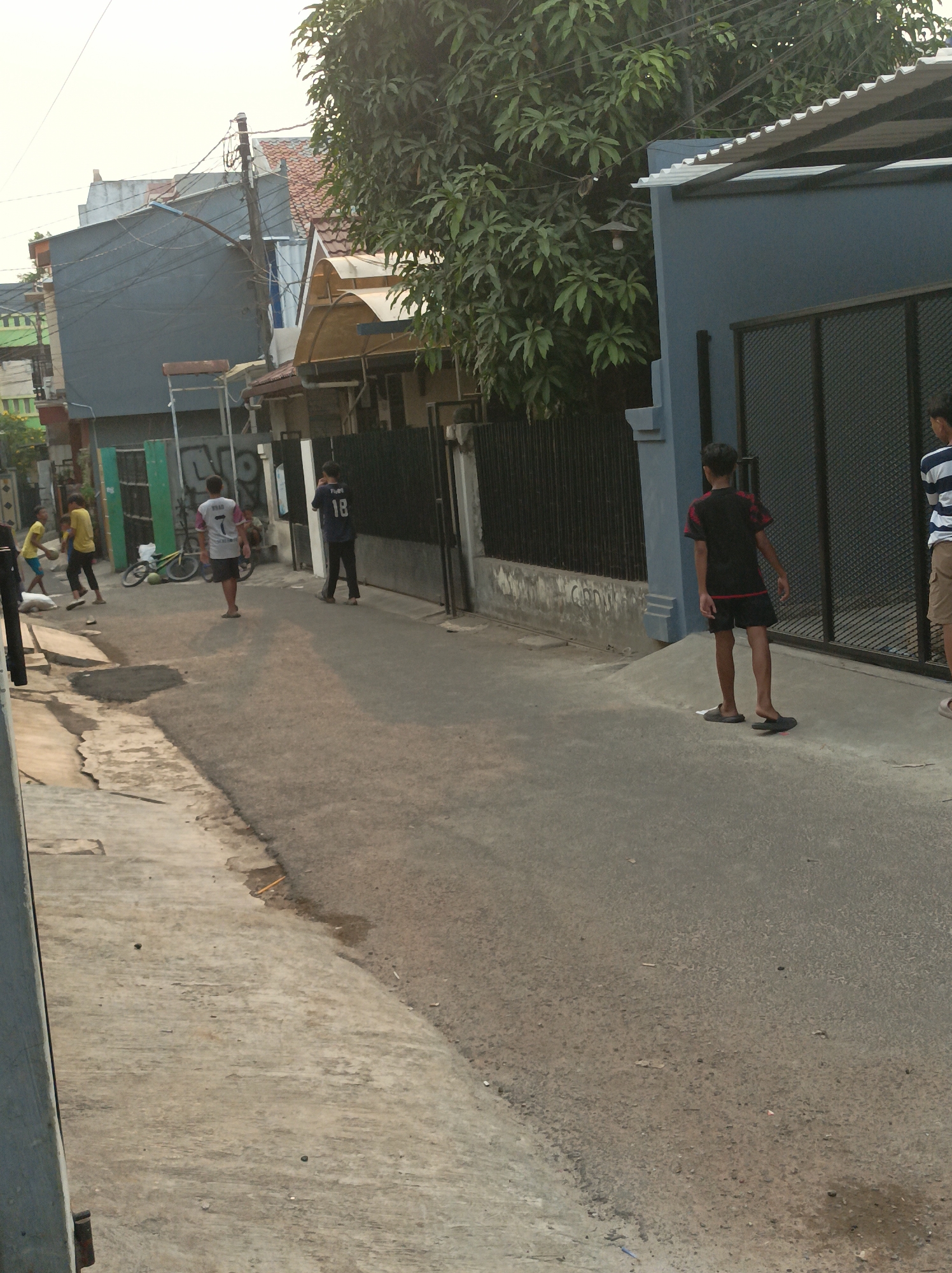The image captures a street scene in West Jakarta, Indonesia, on an overcast day. In the foreground, a concrete pathway runs along the left side of the frame, bordered by a paved street. Several individuals are present in the street. On the left, two young boys in yellow shirts are near a bicycle. Further down the street, a person in a white shirt with a number "7" on the back stands facing away from the camera. To their right, another individual is wearing a dark blue shirt with the number "18". On the far right, a person in a black t-shirt and shorts stands with their back to the camera, and another person in a striped shirt and shorts is partially visible. The environment includes residential buildings with various roofing materials, electrical wires, and trees with dense foliage. The street appears to be a typical urban residential area.