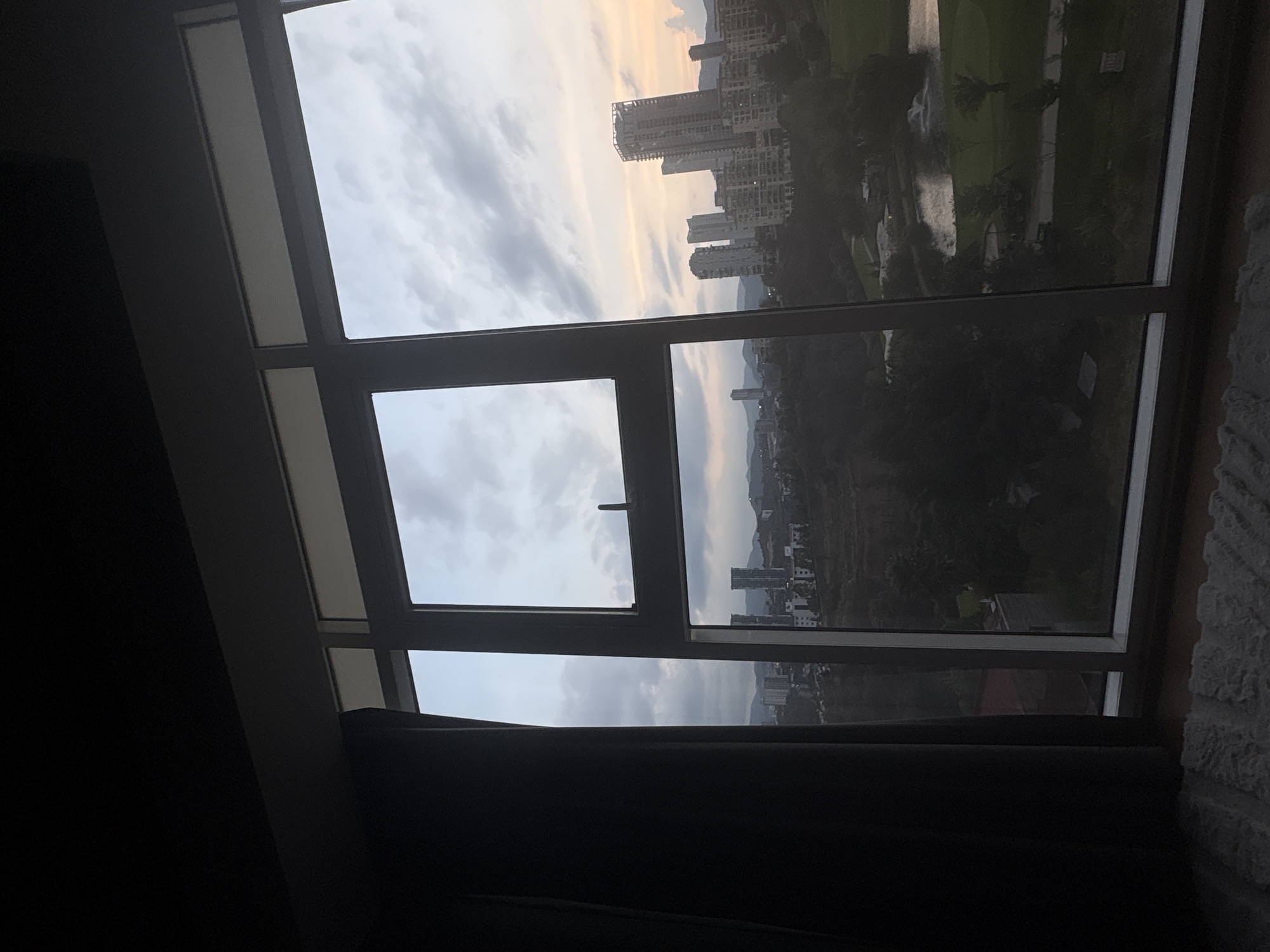 The image presents an interior view of a room looking out through a multi-pane window. The window frames are dark, contrasting with the overcast sky visible through the glass. The view showcases a cityscape in the distance, including high-rise buildings, possibly located in the city of Huixquilucan de Degollado, Mexico. The landscape between the room and the city appears to have trees and green spaces. A curtain is partially drawn on the bottom section of the window, suggesting the room is inhabited. The presence of furniture in the room, and a wall on the right of the frame, indicates the photograph was taken indoors.