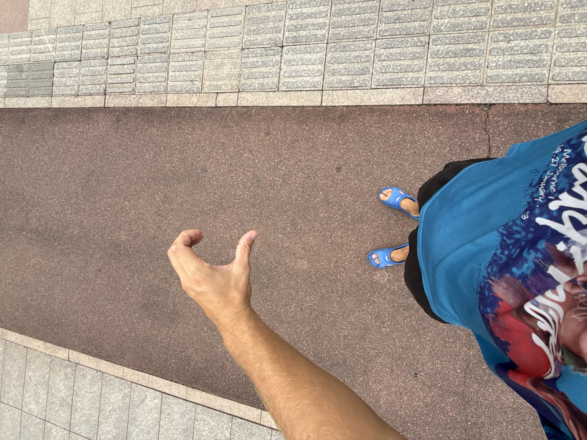 The image shows a high-angle shot of a person's legs and arm, standing on a pavement next to a reddish-brown path. The person is wearing blue flip-flops and a blue t-shirt with a graphic print. Part of the t-shirt has text that appears to read "Melbourne i" and "04-27 January". The person's right arm is extended towards the left of the frame, with their hand held in a cupped shape. The setting appears to be an outdoor urban environment, possibly a sidewalk or park path, with textured paving stones to the left and a darker, granular surface to the right. The lighting suggests it could be daytime, with the sun casting subtle shadows. The overall mood is casual and observational, as if capturing a moment while walking. The location context of Tianhe District, China, is provided as an external detail.