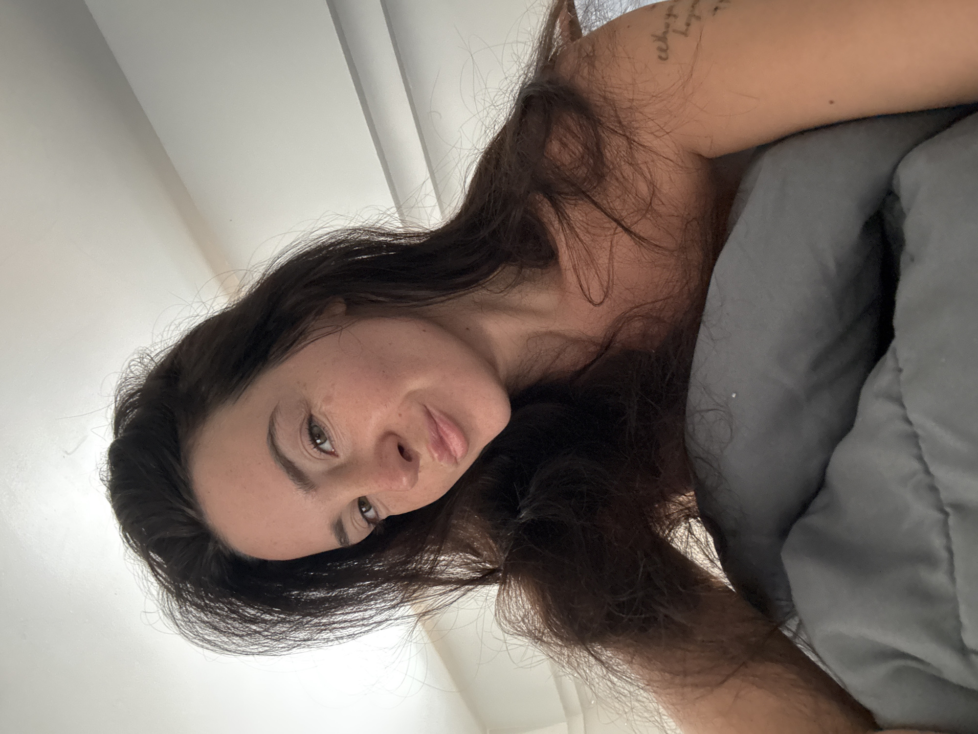A medium-shot selfie shows a woman with long, dark, wavy hair lying on her side in bed. Her face is turned towards the camera, with her head resting on a gray pillow. She has light brown eyes, a small nose, and full lips. Her skin is fair with freckles visible on her cheeks and nose. She is wearing a gray shirt, and a tattoo is visible on her upper arm. The background is a white wall with horizontal molding. The image appears to be taken in North Kuta, Indonesia.