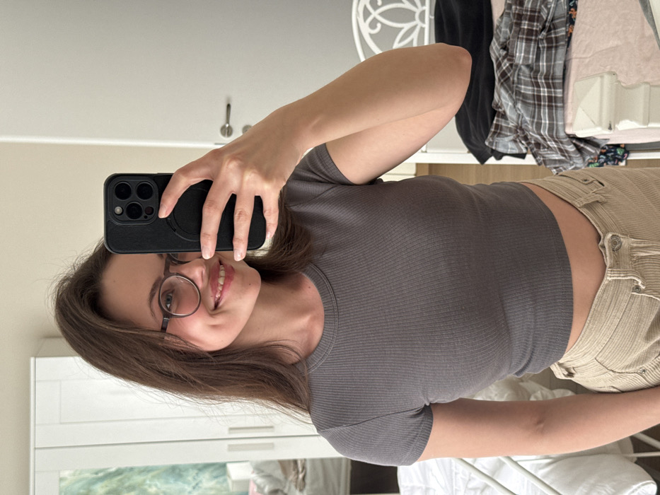 The image displays a female subject taking a mirror selfie indoors. The specific location within the city of Scafati, Italy, cannot be ascertained from the visual data. The female, with brown hair, wears dark-framed round glasses, a dark grey ribbed short-sleeved crop top, and light-colored corduroy pants. She is smiling, revealing teeth and bilateral dimples. Her right hand holds a dark-colored smartphone, encased in black with a circular magnetic attachment, positioned to capture her reflection. The background includes a white two-door wardrobe on the left. To the right, a white metal bed frame or headboard is partially visible, with a plaid shirt and other clothing items casually placed on an adjacent surface. A light-colored door with a handle is visible on the far right. The scene is illuminated by ambient indoor light.