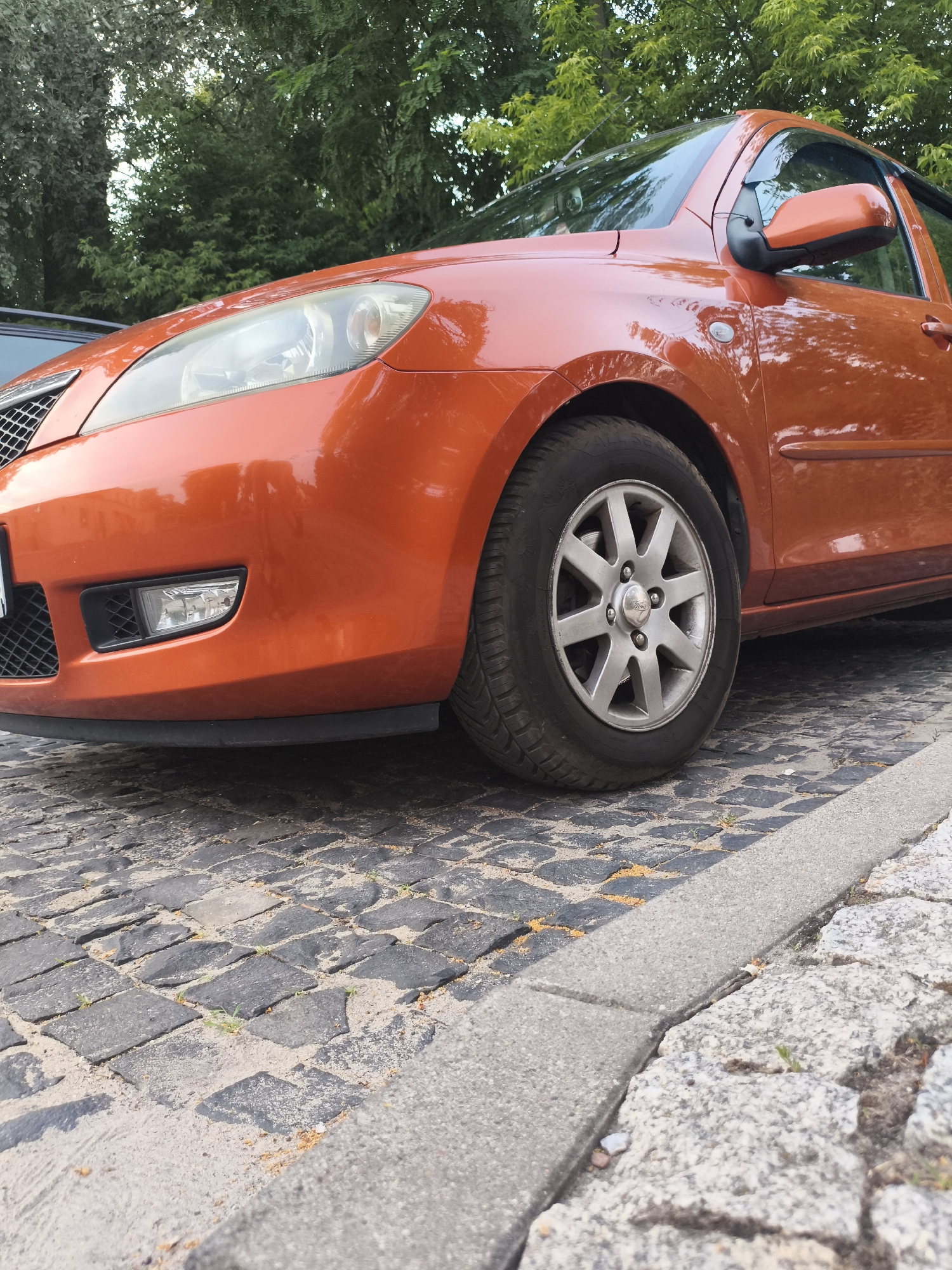 The image shows the front passenger side of an orange car parked on a cobblestone street. The car's headlight, fog light, and a portion of the grille are visible, along with the front wheel and fender. The car appears to be a hatchback, possibly a Mazda. The cobblestone surface is interspersed with sand and small patches of greenery, suggesting an outdoor setting, likely a street or parking area. Trees with lush green foliage form the background, indicating it is likely daytime and possibly spring or summer. The lighting suggests it is not harsh midday sun, but rather a softer light, perhaps late afternoon or early morning. There is no text visible in the image. The setting appears to be a quiet, possibly residential, area in Nowy Dwór Mazowiecki, Poland.