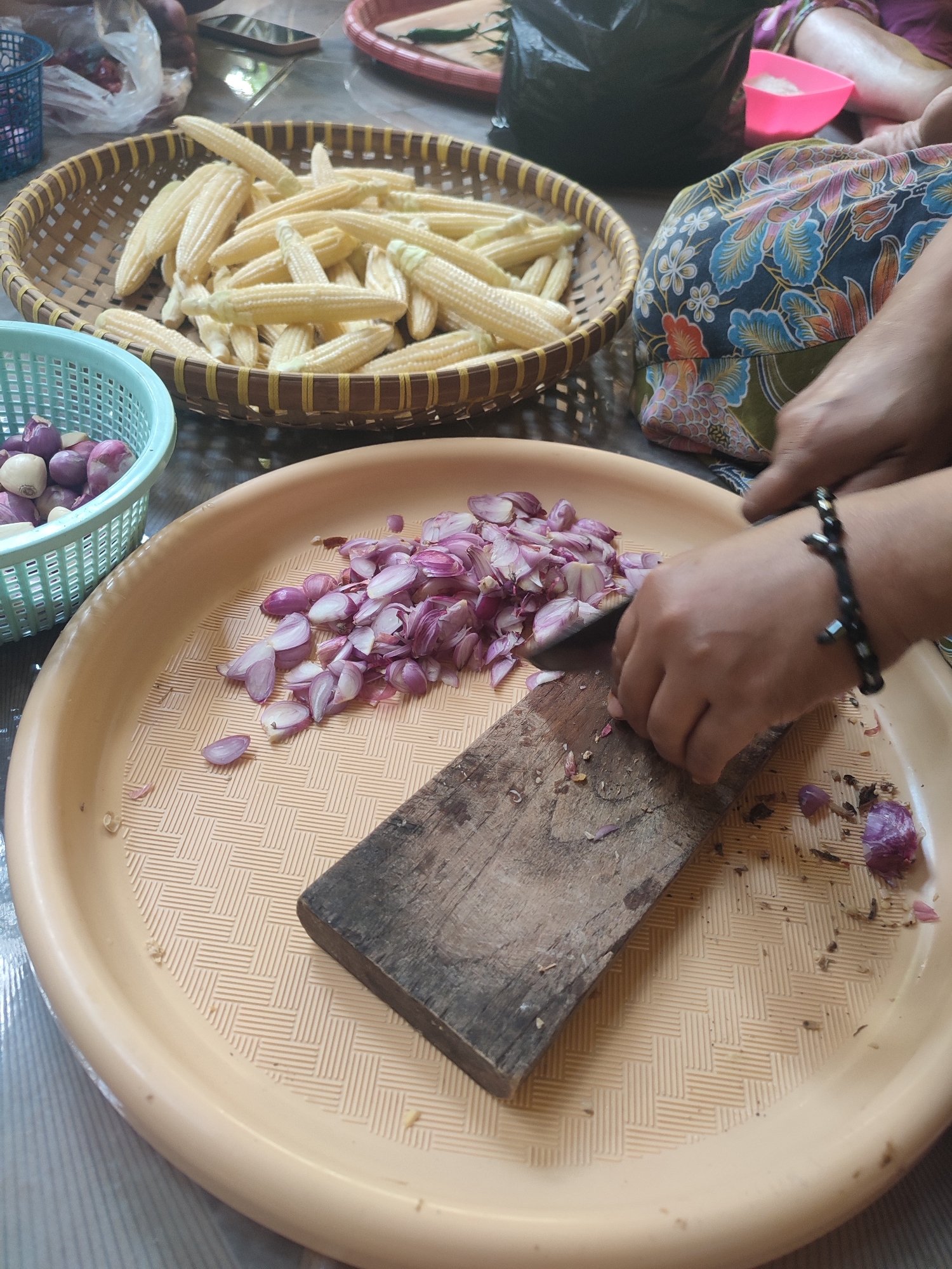 The image captures a close-up of a food preparation scene in Sarwadadi, Indonesia, where a person's hands are actively chopping shallots on a wooden cutting board placed on a beige plastic tray. The tray has a textured surface with a repeating pattern. Sliced shallots are scattered on the tray, along with some whole shallots and discarded skins. To the left of the tray, a light blue plastic basket holds more whole shallots. In the background, a woven basket filled with baby corn is visible. Other elements in the background suggest a domestic or outdoor setting, including a patterned fabric, a black bag, and parts of other people. The lighting is natural, indicating daytime.