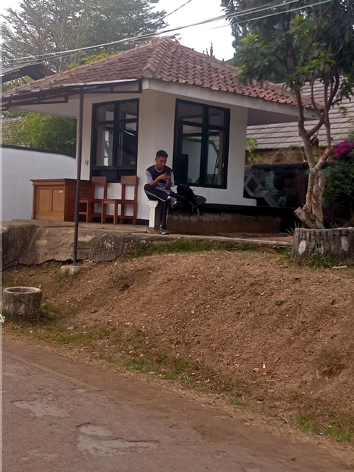 A young man is sitting on a raised concrete ledge in front of a small white building with a tiled roof. He is wearing a dark t-shirt with a lighter gray collar and dark pants. The man is holding a mobile phone in his hands and appears to be looking at it. To his left, there are two wooden chairs and a wooden cabinet. The building has large windows. The scene is outdoors, with dry grass and dirt covering the ground in the foreground and midground. A tree with green leaves is visible to the right of the building. A dirt road with some puddles is in the lower left portion of the image. The image appears to be taken in Sumedang, Indonesia.