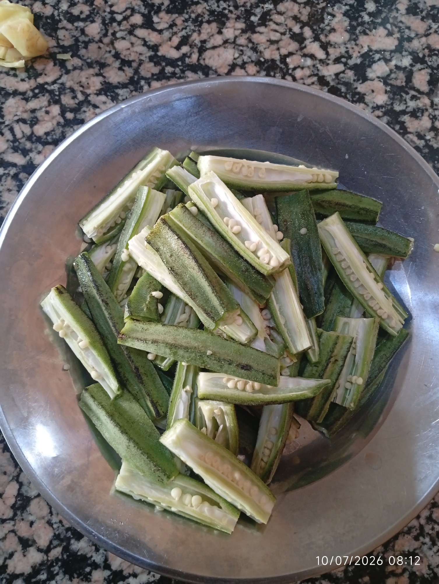 A metal bowl filled with sliced okra is placed on a speckled countertop. The okra pieces are mostly green with white seeds visible inside their hollowed-out centers. Some pieces show evidence of browning. To the upper left of the bowl, there are a few pieces of what appears to be ginger or another root vegetable. The timestamp "10/07/2026 08:12" is visible in the lower right corner of the image. This scene is located in Bana Stream, Nigeria.