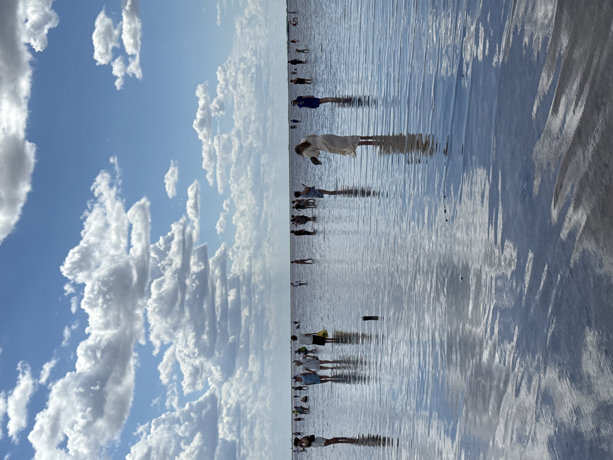 A wide-angle, eye-level shot shows a salt lake with people wading in shallow water. The sky is blue with scattered white clouds. The water is calm and reflects the sky and clouds. A number of people are visible in the distance, scattered across the lake, some walking and some standing. A woman in a light-colored dress stands in the foreground, facing away from the camera. She appears to be holding something in her left hand. In the middle ground, a group of people are gathered, some of them interacting with each other. The overall impression is one of a serene, natural environment being enjoyed by visitors. The location is identified as the Salt Lake in Bursol', Russia.