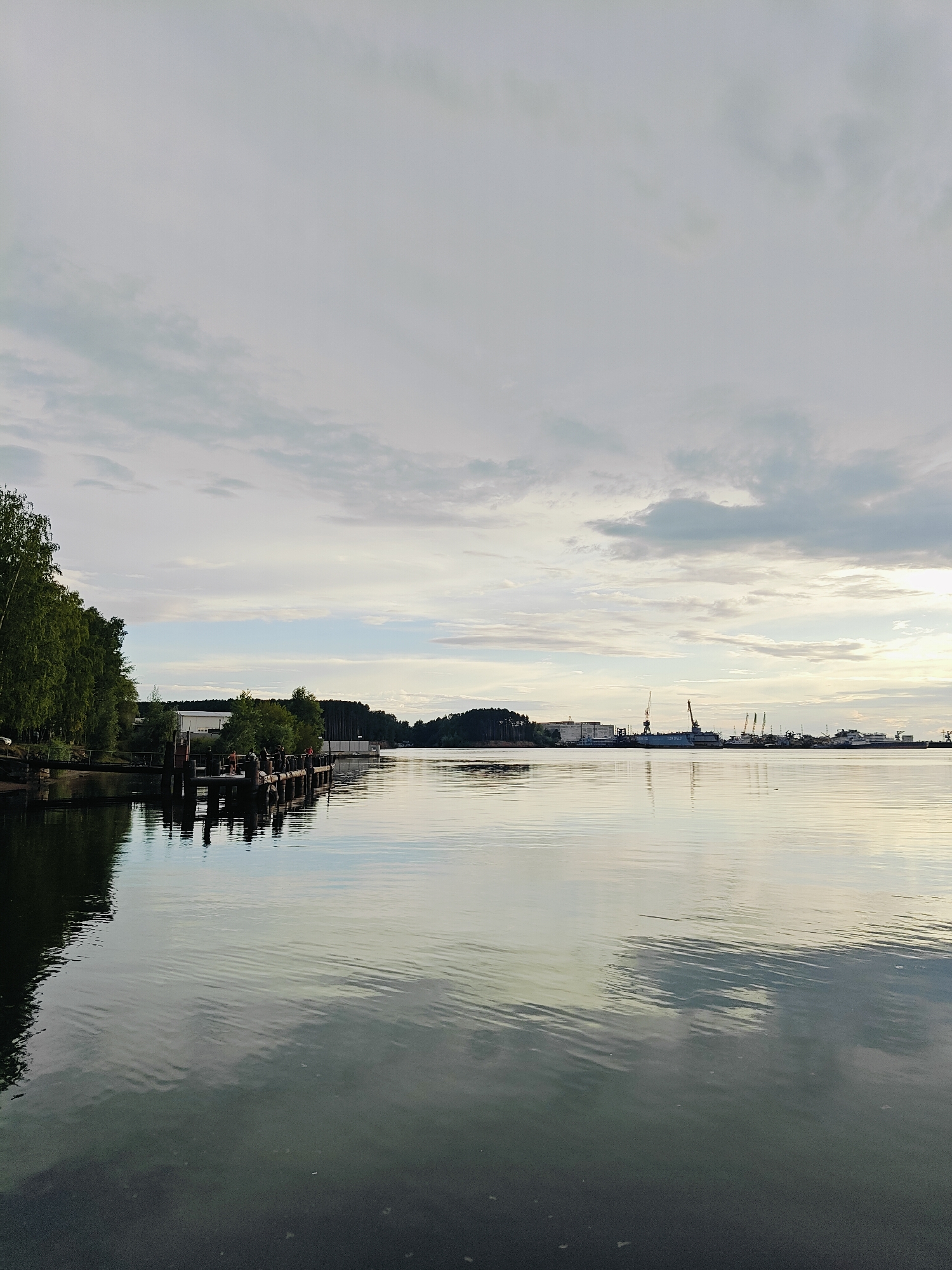 The image depicts a wide body of water, likely a river or bay, under an overcast sky with scattered clouds. On the left side of the frame, a dense line of trees with lush green foliage runs along the bank, reflecting in the water. A wooden pier extends into the water, with several posts and a small structure visible. There appear to be a few people on the pier, though their features are indistinct. Further in the distance, along the opposite bank, a shipyard or industrial area is visible, characterized by several cranes, buildings, and ships. The water is relatively calm, with gentle ripples creating reflections of the sky and the surrounding landscape. The overall impression is one of a serene, industrial waterfront scene. The location in the city of Chaikovsky, Russia, is consistent with the presence of a large river (Kama River) and industrial activity, particularly shipyards.