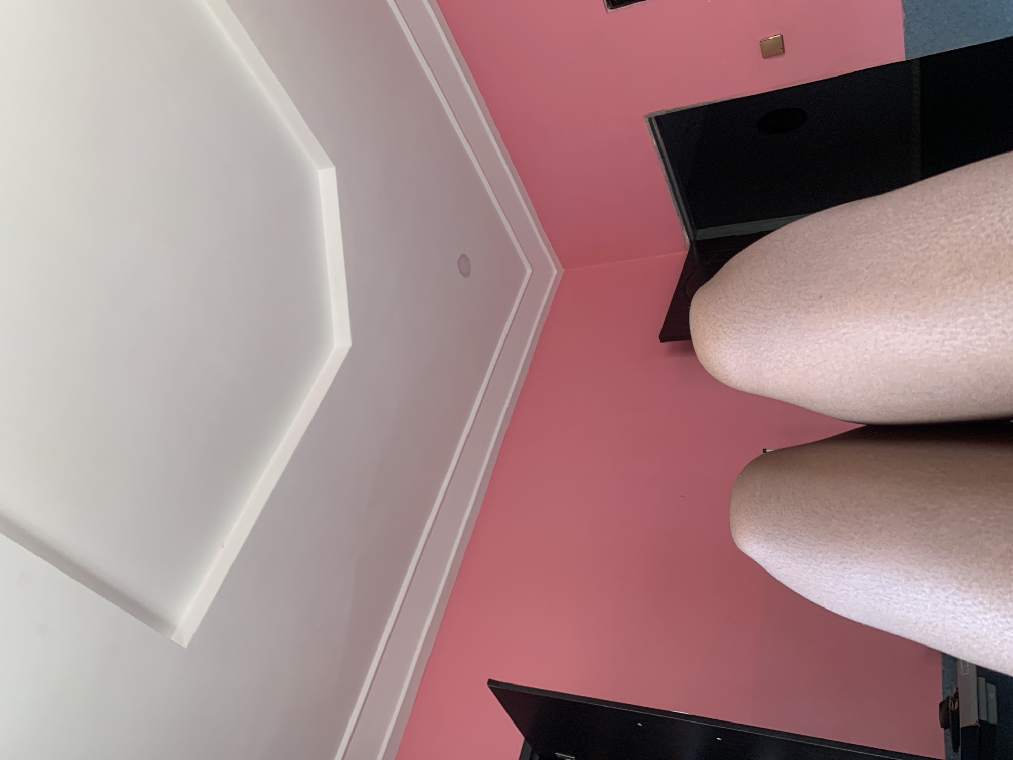 The image shows a low-angle view of a room. The walls are painted a bright pink, and the ceiling is white with decorative molding. In the foreground, the tops of two legs are visible, appearing to belong to a person lying down or sitting. The skin texture on the legs is clearly visible. There are two dark doorways on the right side of the image, one of which is partially open, revealing darkness within. A light switch or electrical outlet is visible on the pink wall near one of the doorways. A black object, possibly a cabinet or furniture, is on the left side of the image. The scene appears to be indoors, likely a residential bedroom or living space. The location is specified as Kakau, Nigeria.