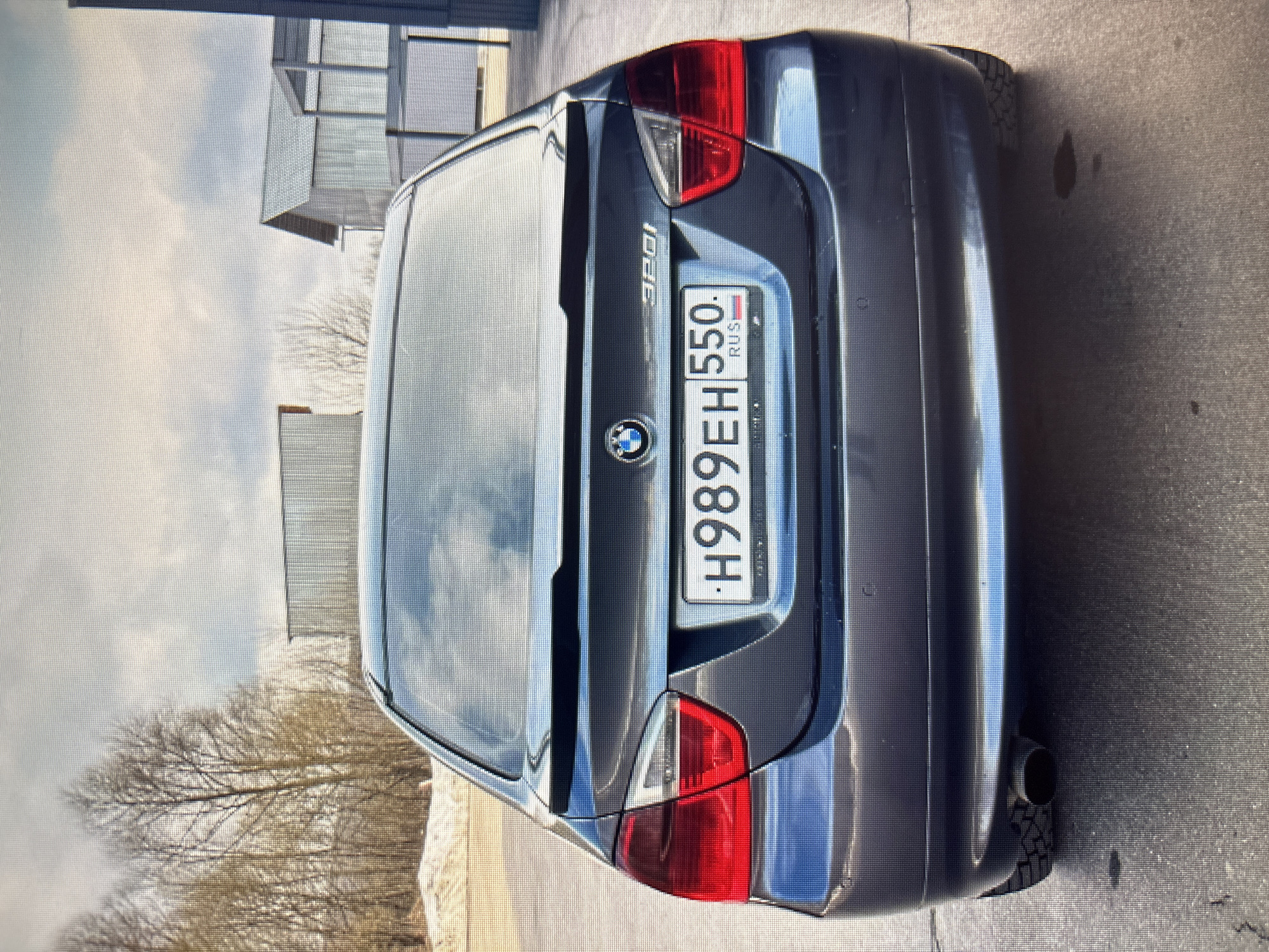 The image shows the rear view of a dark grey BMW 320i sedan parked on an asphalt surface. The car has a license plate with the inscription "H989EH 550. RUS". In the background, there are bare trees, some snow patches, and modern-looking buildings with grey facades and pitched roofs. The sky is overcast with clouds. The scene suggests an outdoor setting in a location that appears to be a semi-urban or industrial area. There are no people visible in the image. The focus is on the car and its immediate surroundings.