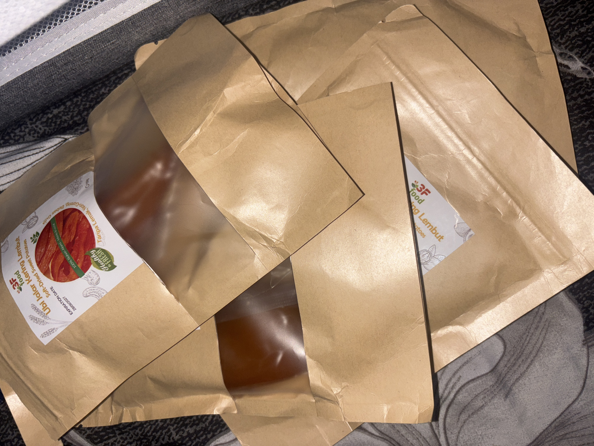 A close-up, overhead shot shows several brown paper pouches with clear windows, stacked on a dark, textured fabric surface. One pouch prominently displays a label with the text "3F Food Ubi Jalar Kering Lembut Soft-Dried Sweet Potatoes" and an expiration date of "20/06/2027". The label also features a graphic of sliced sweet potatoes and a "Healthy & Fresh" logo. The contents of the pouches appear to be a reddish-brown, solid food item. The scene is indoors, and the lighting suggests it might be artificial. There is no information or visual evidence to suggest the location is in West Jakarta, Indonesia.