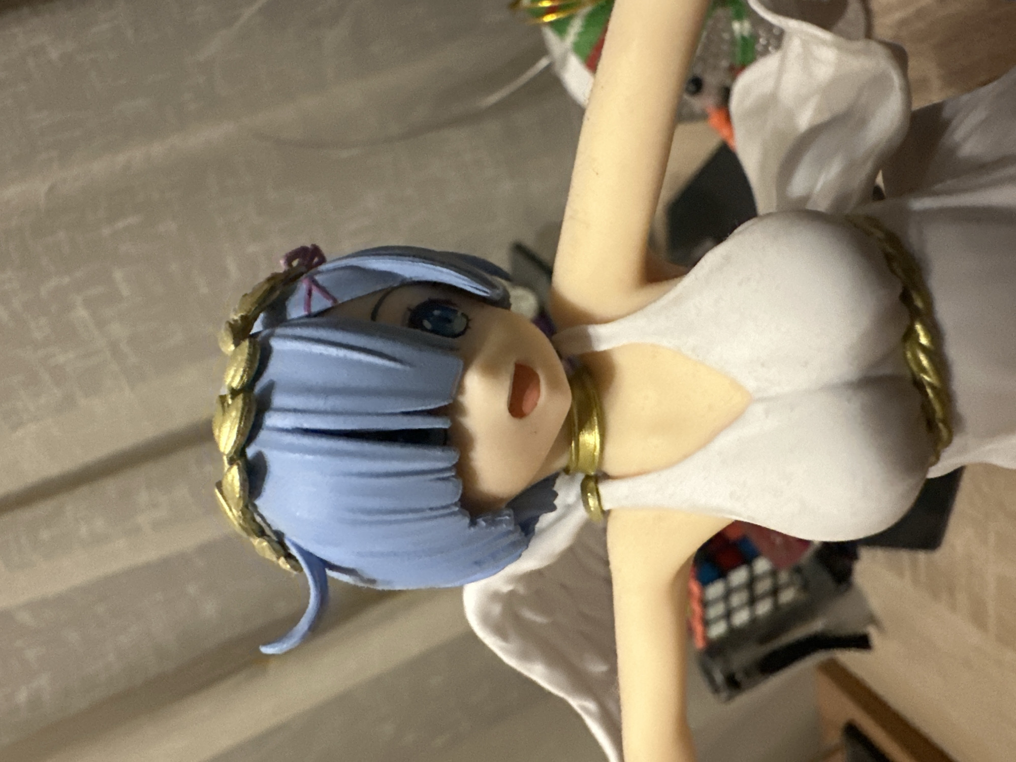 A close-up, eye-level, slightly high-angle shot shows a detailed anime figurine of a character named Rem from the anime series Re:Zero. The figurine is depicted as an angel, with light blue hair styled in a bob with bangs covering her eyes and a gold laurel wreath adorning her head. She has blue eyes, visible beneath her bangs, and a small, open mouth. Rem wears a white, sleeveless dress with a gold choker around her neck and a gold belt tied around her waist. White, feathery wings are attached to her back. The background consists of a textured, beige curtain. To the left of the figurine, partially visible, are a black electronic device with colorful buttons and a wooden surface. To the right, out of focus, are colorful elements, possibly part of another decoration or figurine. The lighting is even, highlighting the detail of the figurine. The figurine is positioned centrally in the frame, with its arms extended slightly outwards. The setting is indoors, with no discernible urban or natural landmarks.