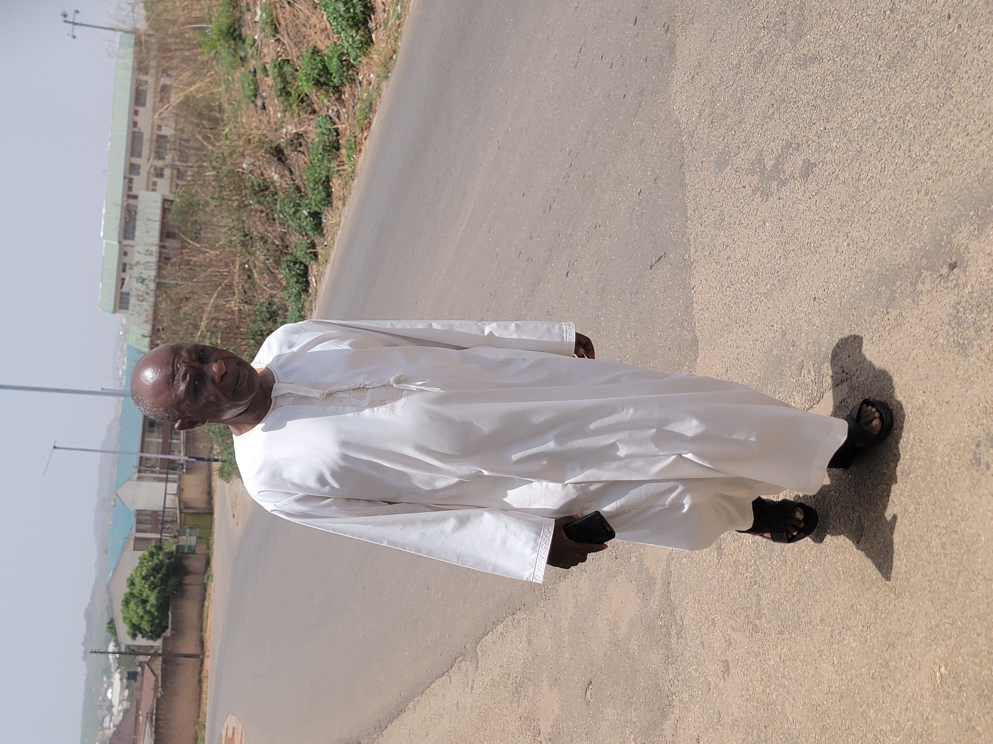 A middle-aged man of African descent is standing on a paved road. He is wearing a long white garment and black sandals. The background includes a building complex, vegetation, and the sky. The location is likely in Jos, Nigeria.