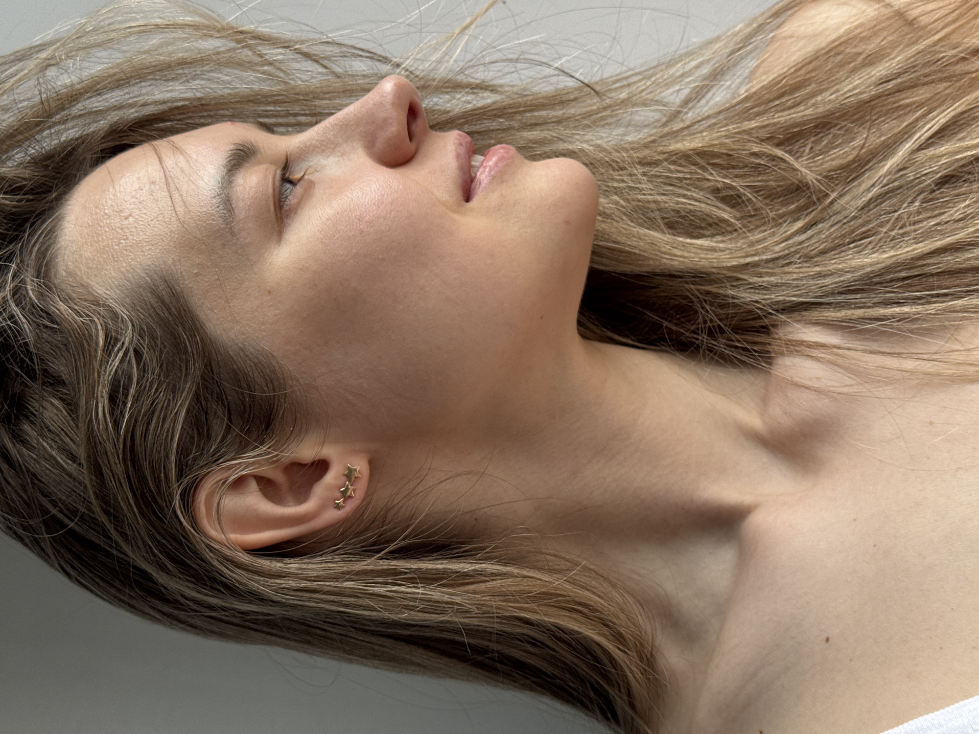 A woman with light brown hair lies down, facing upwards and slightly to the right. Her face is pale and her lips are slightly parted. She has small gold star earrings in her left ear. Her neck is slender. She appears to be in an indoor setting. The location is not discernible from the image.