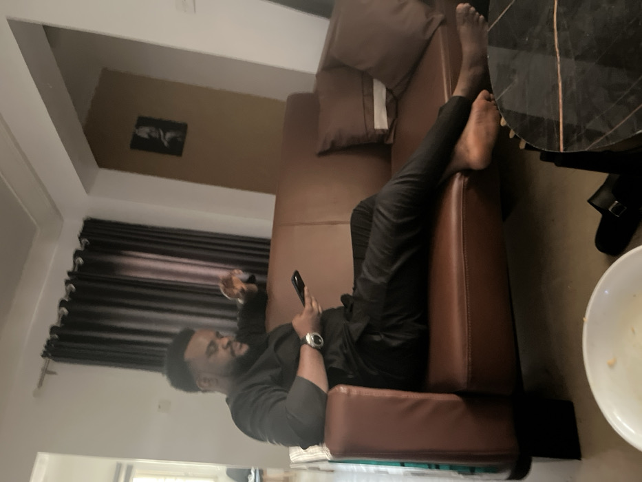 The image displays an indoor scene, likely a living room in Kurudu, Nigeria. A man with a beard is relaxing on a brown, leather-like sofa. He is dressed in a dark long-sleeved top and dark trousers, with a metallic watch on his left wrist.
The man is lying on his back, looking at a smartphone held in his left hand. His right hand appears to be holding a small, dark object, possibly a remote or a snack. His bare feet are visible, resting on the sofa's armrest or backrest on the right side of the frame. Brown cushions are placed on the sofa.
In the foreground, to the right of the sofa, a dark, possibly marbled, round table is partially visible. Below the table, a white bowl containing some food residue is present, along with a dark slipper or sandal on the floor.
The background features dark horizontal blinds or curtains covering a window on the left. Above the curtains, a light-colored wall holds a small, framed black and white picture. The ceiling is also visible, completing the indoor setting. The scene conveys a sense of leisure and comfort. No visible text is discernible in the image.