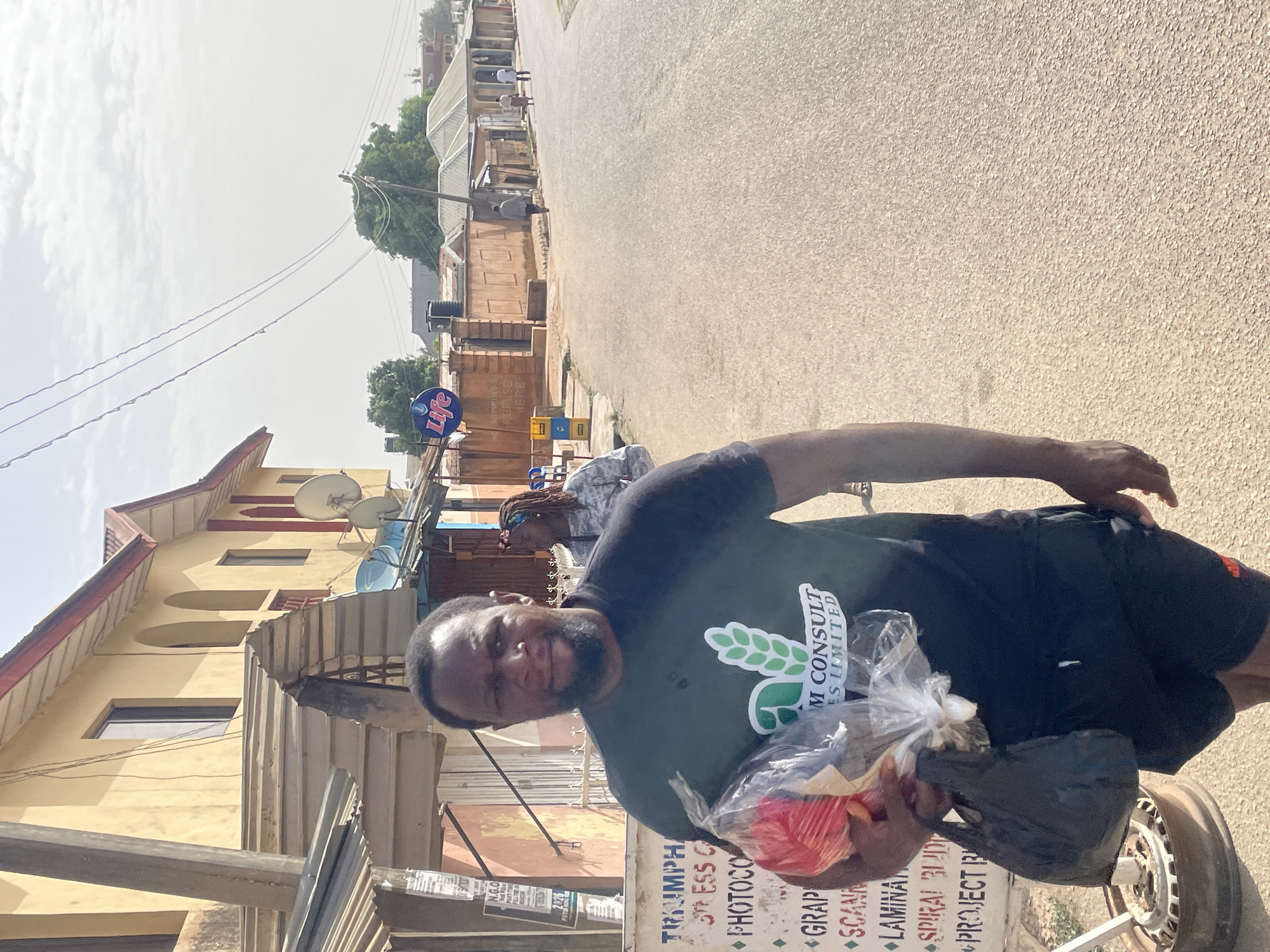 A man is standing on a street in Jos, Nigeria. He is wearing a black t-shirt with the logo of "M Consult Es Limited" on it, and black shorts. He is holding a plastic bag containing red objects and a black bag. Behind him, a woman with dreadlocks is visible.
The street is lined with buildings, some of which are yellow with red trim. A sign for "Life" is visible in the background. Additionally, a sign for a business named "TRIUMPH" offering services such as photocopying, scanning, and lamination can be seen in the foreground. Overhead, there are power lines. The street appears to be paved.