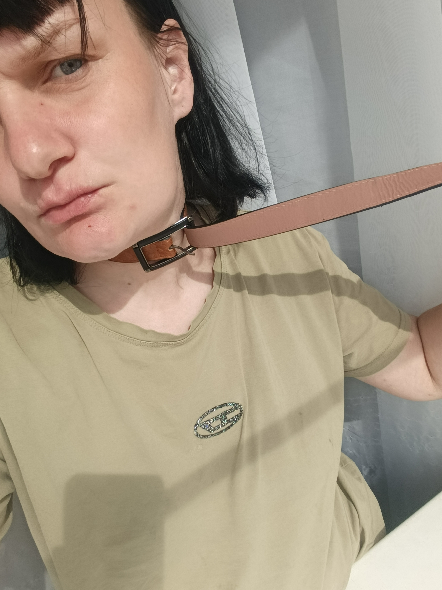 A chest-up portrait captures a woman indoors, possibly in Фоки, Russia. She has dark, chin-length hair with bangs framing her forehead, and striking light blue eyes that gaze directly at the viewer. Her expression is neutral, with her lips slightly pursed.
She is wearing a simple khaki or olive green short-sleeved t-shirt, which features a shimmering, textured, stylized logo on the left side of the chest. Around her neck, a light brown leather belt is buckled, functioning as a choker. The silver buckle and the leather strap are prominent at the base of her throat. The longer end of the belt extends out of the frame to the right, held by her hand, giving the impression of a leash.
The background consists of light-colored, possibly white or pale grey, draped fabric or a plain wall, creating a soft and uncluttered setting. The lighting is even and soft, suggesting an indoor environment without harsh shadows. No specific time of day or weather conditions are discernible from the image. The logo on her shirt is stylized and not clearly legible as text.