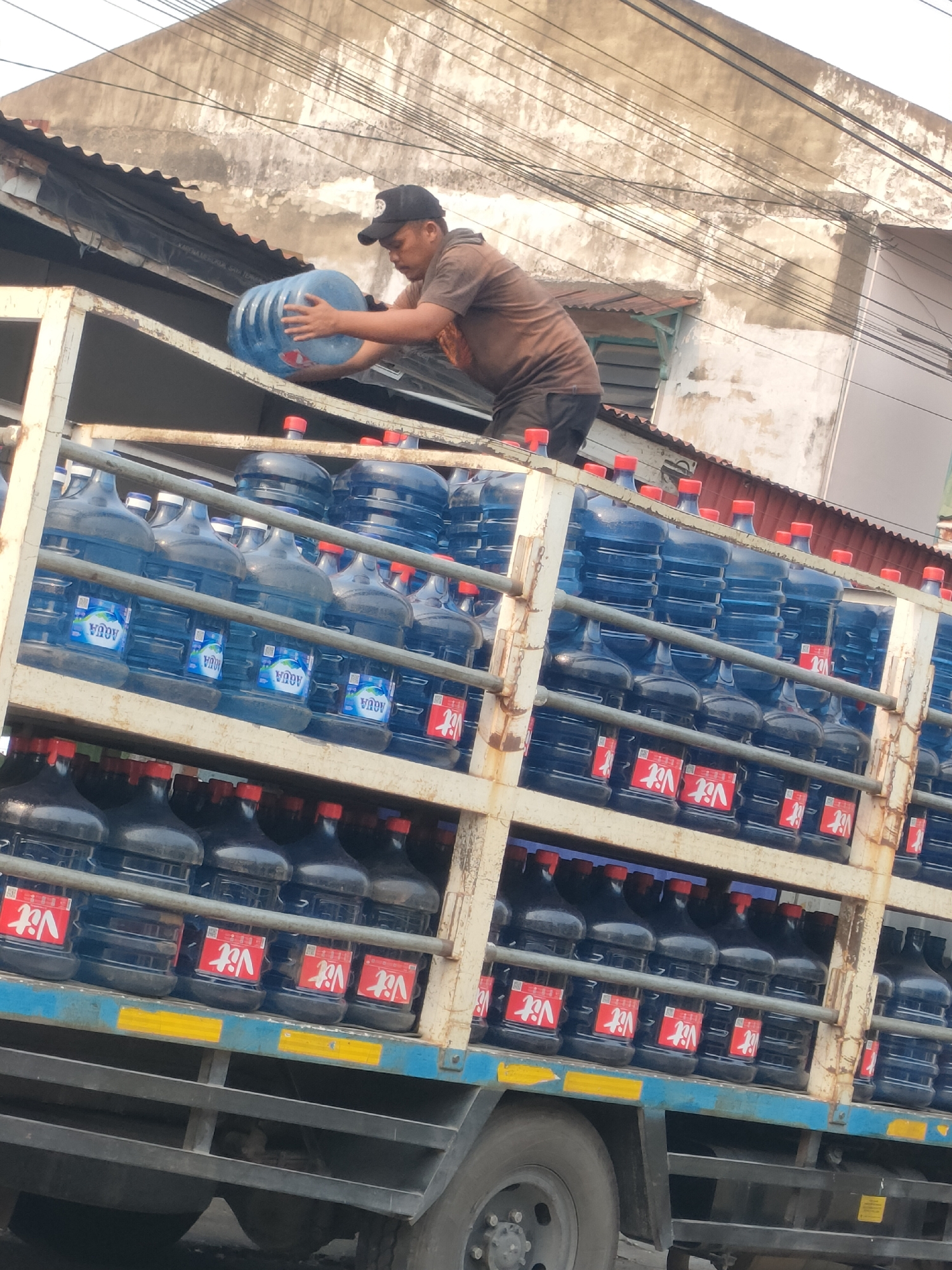 A man wearing a black baseball cap and a brown t-shirt is seen on top of a truck, holding a large blue water jug. He is in the process of placing it onto the truck bed which is filled with many similar blue water jugs with red caps. The jugs are stacked on multiple shelves. The truck appears to be parked in an urban environment with buildings visible in the background. The scene is likely in Pekanbaru, Indonesia, given the context.