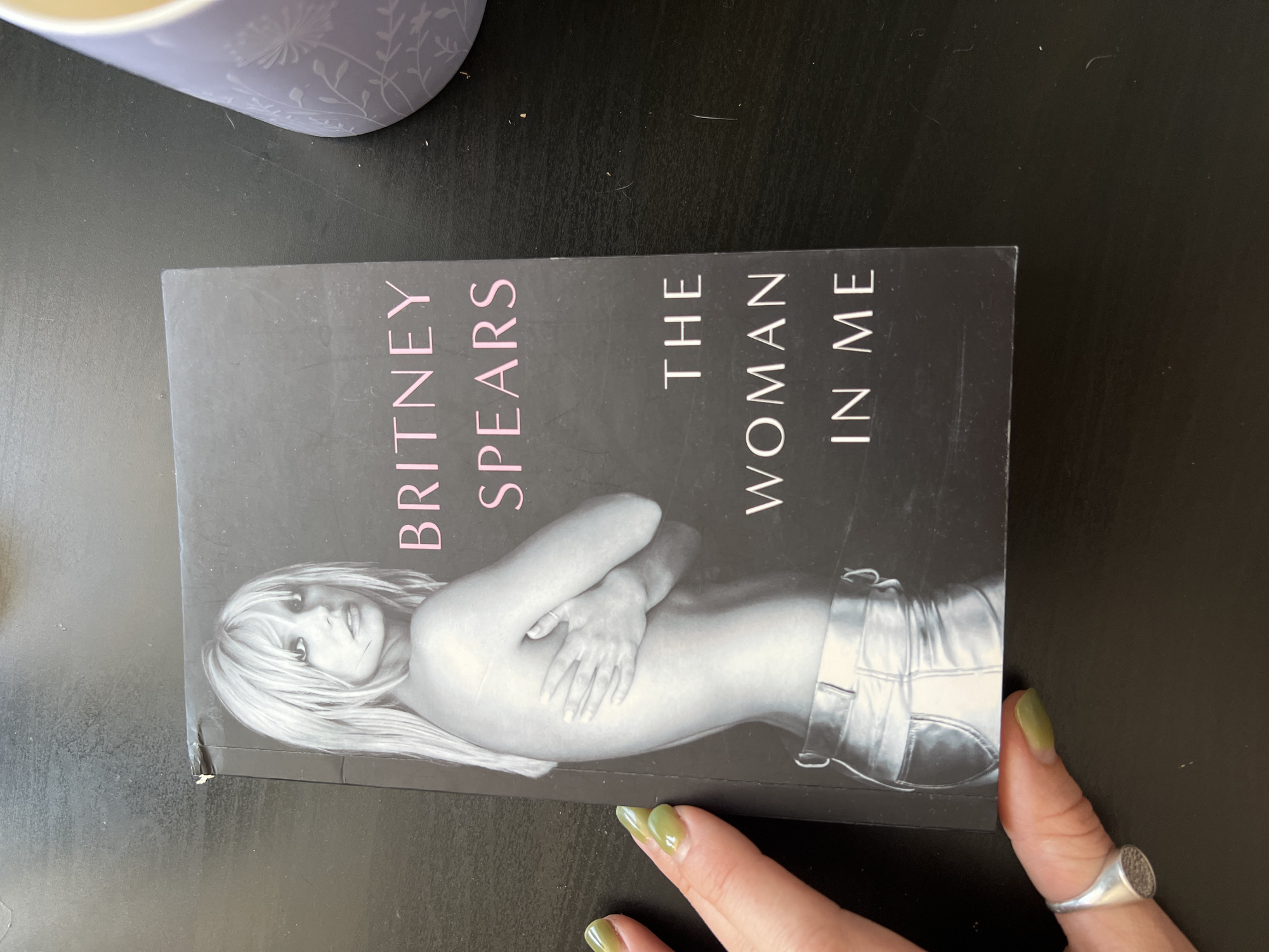 An open book titled "The Woman In Me" by Britney Spears is on a dark, flat surface, likely a table. A person's left hand, with light green nail polish and a silver ring on the index finger, is visible on the lower left side of the image, holding down the book. To the right of the book, a portion of a light purple mug with a floral pattern is visible. The book's cover features a black and white image of Britney Spears and the title text is in pink and white. There is no information in the image to determine its location in Novosibirsk, Russia.