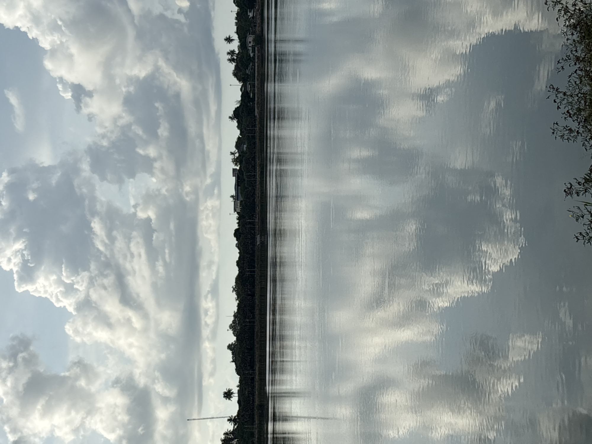 The image shows a long body of water, likely a river or canal, reflecting the sky, which is filled with clouds. A line of trees and buildings runs along the edge of the water, indicating a populated area. Palm trees are visible among the other trees. The location is บ้านบางรักษ์, Thailand.