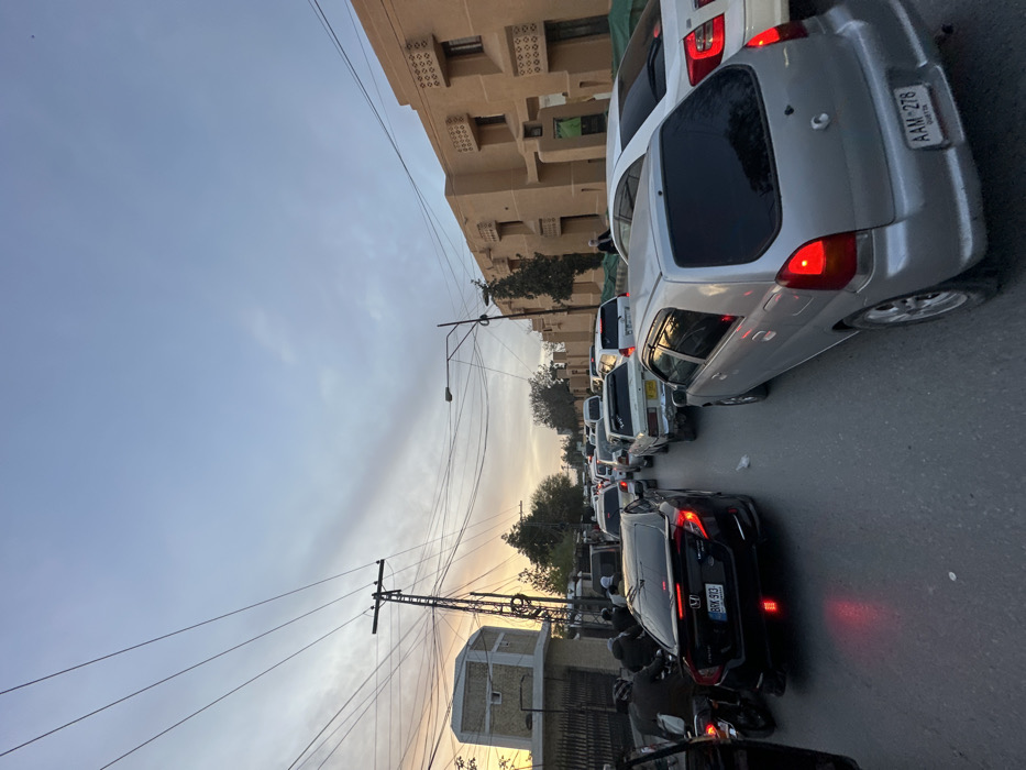 The image depicts an urban street in Quetta City Tehsil, Pakistan, at dusk, characterized by heavy traffic congestion. The sky exhibits a gradient from orange and pink near the horizon to light blue overhead.
In the foreground, a silver Suzuki Alto hatchback (license plate AAM-278, Quetta) is positioned on the right side of the frame, with its red brake lights illuminated, indicating a stopped or slow-moving state. To its left, partially obscured, is a white sedan. On the left side of the frame, the rear of a black Honda Civic sedan (license plate BRK 913) is visible, also with illuminated red brake lights. Behind the Honda Civic, several motorcycles with riders are stopped.
The asphalt road surface shows multiple lanes of vehicles extending into the distant background, all appearing to be stopped or moving slowly. A diverse array of cars and vans, including older white sedans and a white van, populate the roadway.
On the left side of the street, a light-colored, hexagonal-shaped building with a flat roof and small rectangular windows is visible. A prominent black utility pole, laden with numerous overhead power lines, stands between this building and the road.
On the right side of the street, a multi-story building with a light brown facade features distinctive square windows with decorative grilles above them. A street light pole is positioned in front of this building. A section of green netting or tarp partially covers the lower part of the building. One male pedestrian, dressed in light-colored traditional clothing, is visible walking along the sidewalk near this building. Sparse trees are visible in the background between buildings and along the street.