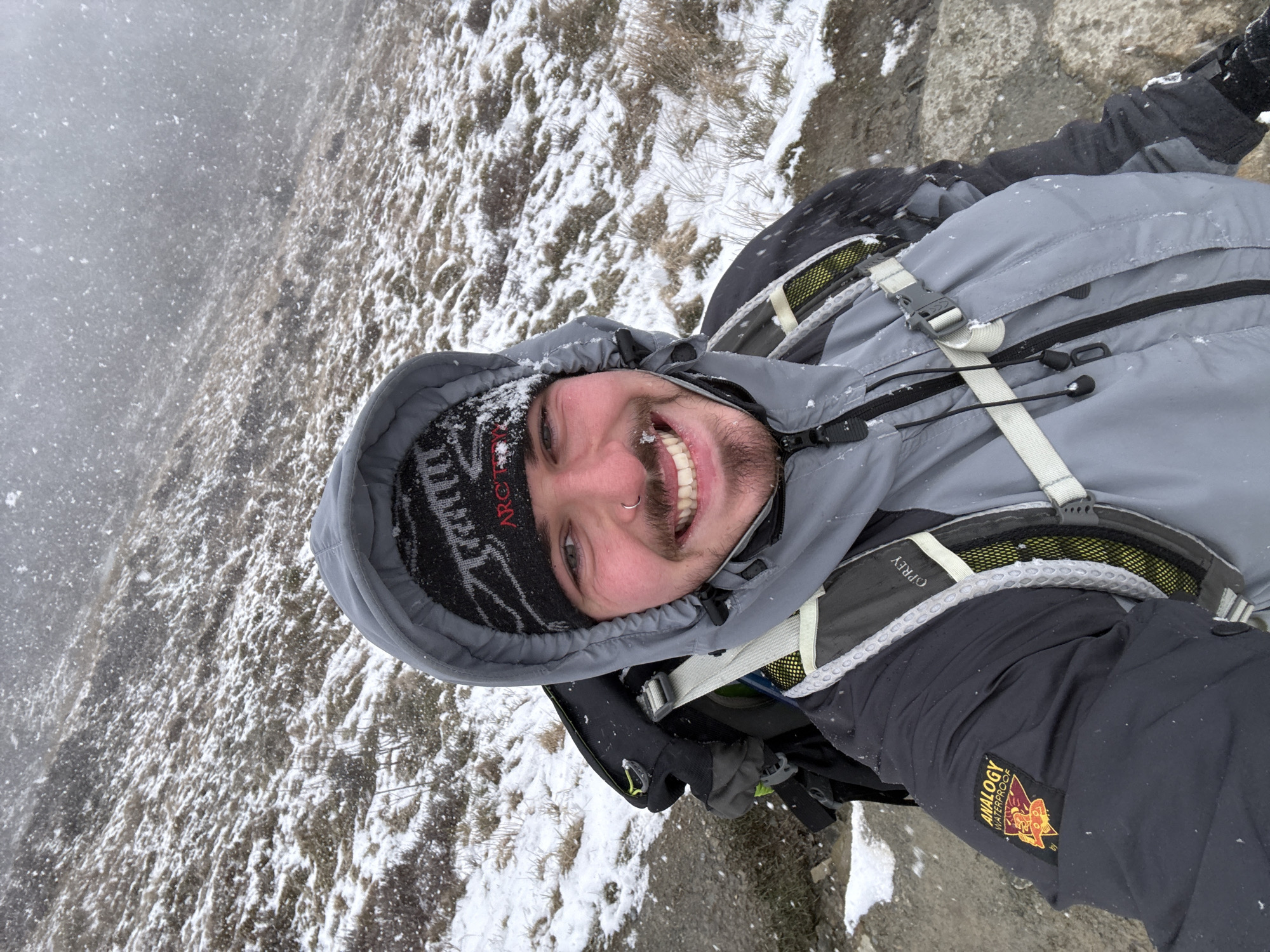 The image displays a selfie of a male individual outdoors in a snowy environment. The subject is a light-skinned male with a short beard and a silver nose ring, smiling directly at the camera. He is wearing a black knit beanie with a grey pattern and red "ARCTERYX" text, a grey hooded jacket, and a dark under-jacket with an "ANALOGY WATERPROOF" patch on the left sleeve. A backpack with visible "OSPREY" branding and light grey straps with green mesh accents is worn on his back. Snowflakes are actively falling and are visible on his clothing, beanie, and facial hair. The background consists of a sloping hillside covered in patches of snow and sparse, dry brown vegetation. A portion of a dirt or rocky path is visible to the left of the subject. The location is an unidentifiable mountainous or hilly region in Unknown, Unknown.