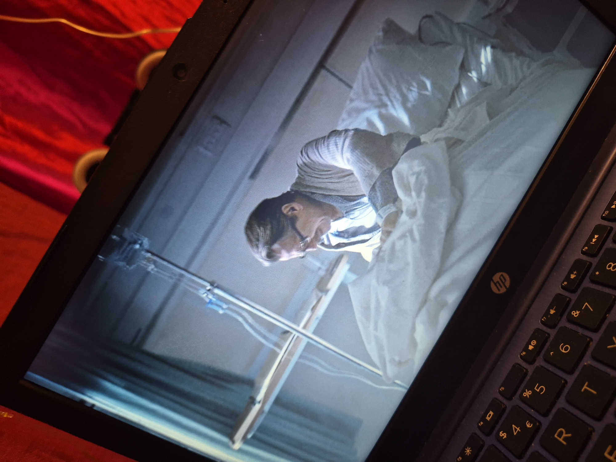 The image shows a laptop open with a movie playing on the screen. The movie appears to be set in a hospital room, with a man lying in a hospital bed. He is wearing glasses and a cardigan over a striped shirt. Medical equipment, including an IV stand, is visible in the background. The laptop keyboard is partially visible in the foreground, with the HP logo on the right side. The background behind the laptop is a red, textured fabric. The scene depicted on the laptop screen is brightly lit, while the foreground and surrounding environment are darker. There is no information within the image to suggest a specific location in Sandwell, United Kingdom.