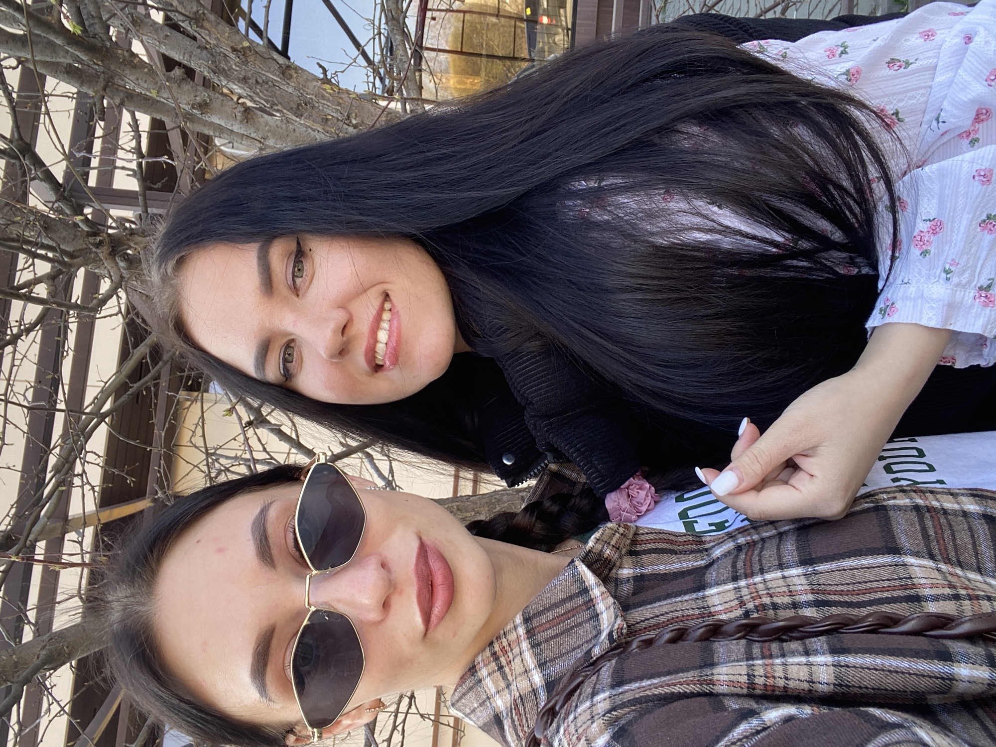 Two women are posing outdoors. The woman in the foreground is wearing sunglasses and a brown plaid shirt. The woman in the background has long, dark hair and is wearing a white floral top. They are possibly located in Chaikovsky, Russia, based on information provided. The background includes tree branches and a glimpse of buildings. The scene appears to be an informal, possibly spontaneous snapshot.