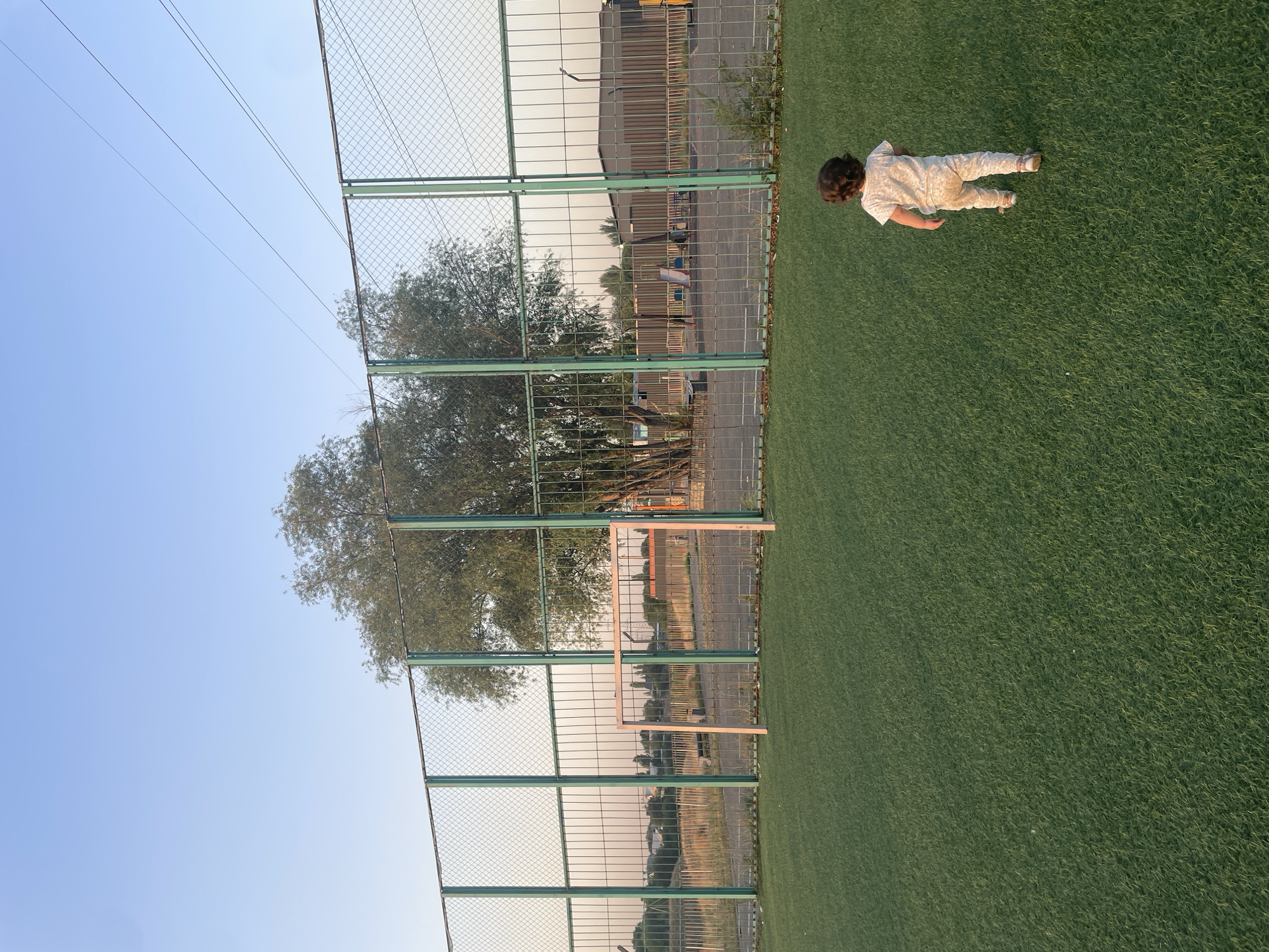 The image depicts an outdoor artificial turf field, likely a sports field, situated in Almaty, Kazakhstan. A young child with curly dark hair, seen from the back, is walking away from the camera across the green turf. The child is wearing light-colored pajamas with a subtle pattern. In the background, a chain-link fence with green metal posts encloses the field. Beyond the fence, a small, light-colored soccer goal is visible. Further back, a large tree with dense foliage stands, and beyond that, there are buildings and vegetation, indicating an urban or semi-urban setting. The sky is a clear, pale blue, suggesting it is either early morning or late afternoon. The lighting indicates the sun is low in the sky, casting soft shadows.