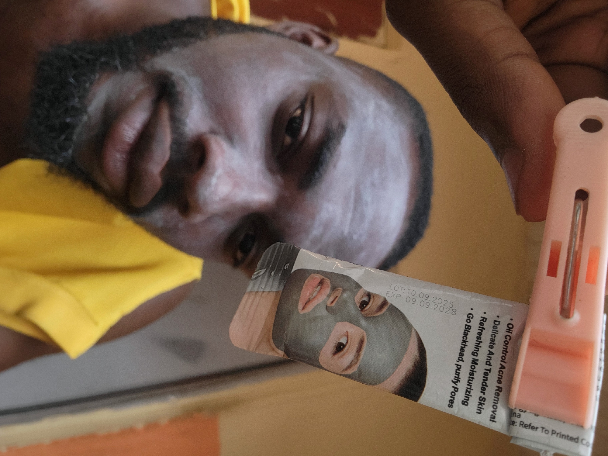 A low-angle shot shows a Black man with a beard and short hair, wearing a yellow shirt. He is holding a sachet of face mask product with a pink plastic clip. The text on the sachet indicates oil control acne removal, delicate and tender skin, refreshing moisturizing, and blackhead purification. The expiration date is 09.09.2028. The location is possibly Jos, Nigeria.
