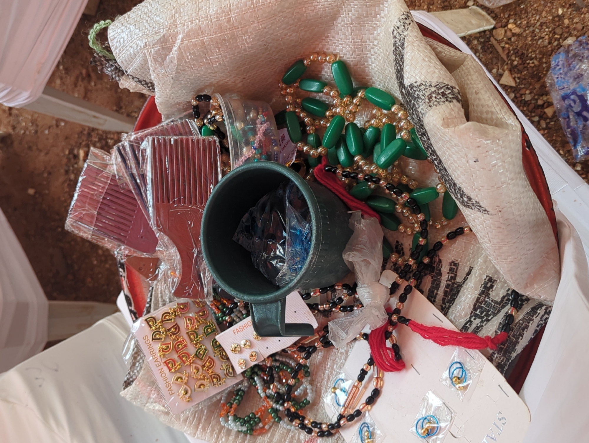 A close-up, overhead view captures a collection of various small goods and accessories contained within a woven bag, likely set in an outdoor or semi-outdoor environment in Unguwar Rimi, Nigeria.
The bag, made of a light-colored, coarsely woven material with dark, patterned sections, is filled with items. Prominently featured are multiple strands of beaded necklaces; one notable type consists of large, oval, green beads interspersed with smaller gold-colored beads. Other necklaces include combinations of black, gold, orange, and multi-colored beads, some tied with a red thread or cord.
To the left, several brown plastic combs are visible, each individually wrapped in clear plastic packaging. A dark green mug sits near the center, also holding some items wrapped in clear plastic. Various earrings are displayed on cardboard backings: one card clearly states "FASHION EARRINGS" and shows numerous small, gold-colored, letter-shaped studs. Another card displays multiple gold stud earrings. Further down, small blue hoop earrings are visible on a card, with the partial word "STA" printed on it, possibly for "Stainless." A small white label with the text "GARANT" and the number "1" is also present, suggesting a guarantee.
The background outside the bag reveals reddish-brown ground, possibly dirt or gravel, and parts of a white fabric structure, which could be a tent or tablecloth. The bright, even lighting suggests it is daytime. No specific activity or individuals are depicted, and no particular emotions are conveyed.