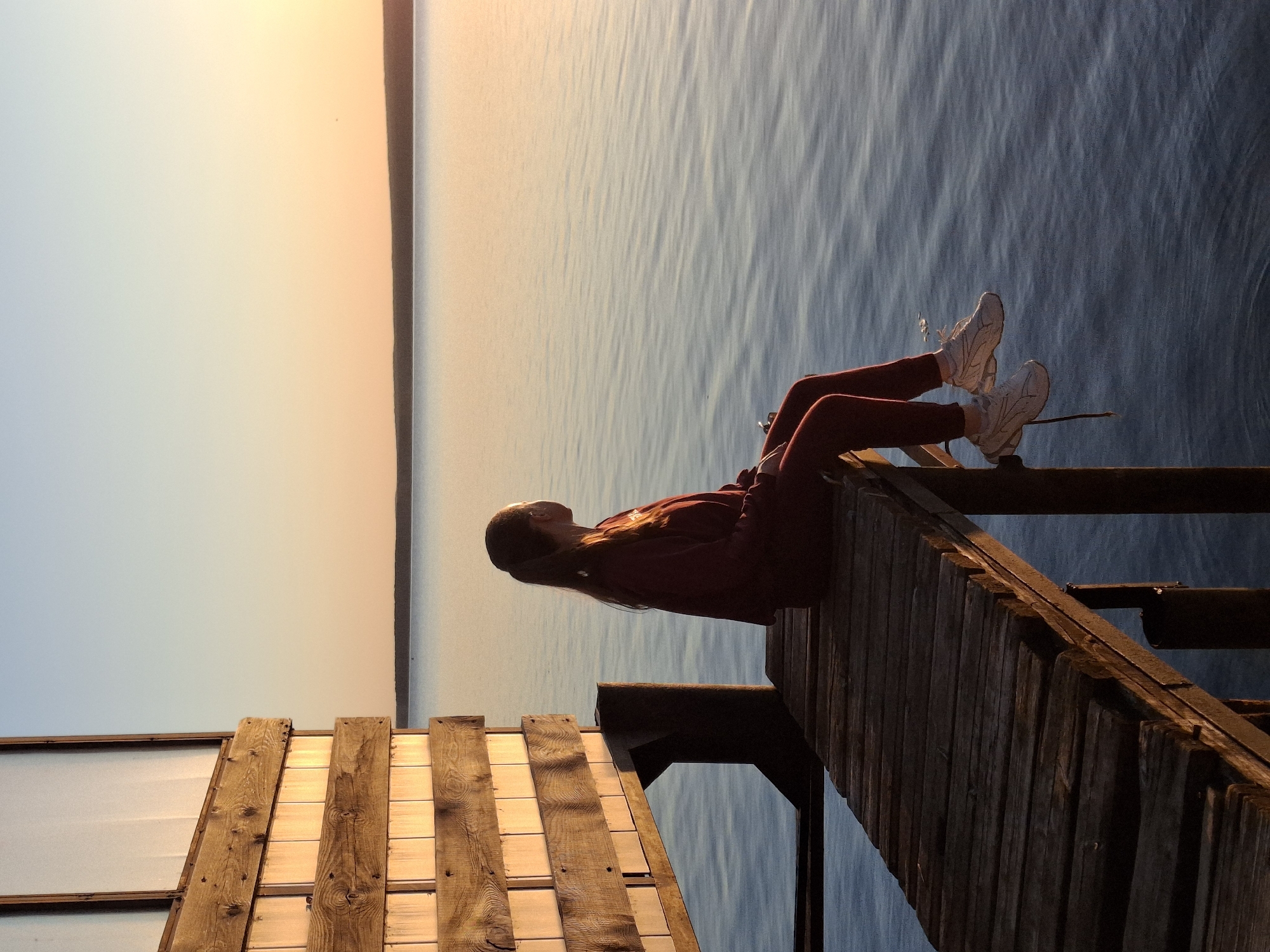 A woman sits on the edge of a wooden dock, feet dangling, overlooking a large body of water under a sky gradient from blue to orange. The scene includes a nearby wooden structure attached to the dock. The location is potentially in Chaikovsky, Russia. The time of day appears to be sunset.