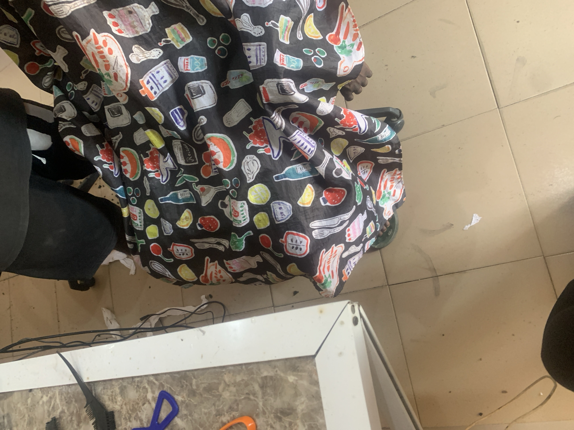 The media file depicts a close-up view of a black cape with a whimsical pattern of food and kitchen items. The cape is draped around a person, whose dark clothing and legs are partially visible, suggesting they are seated or standing. The pattern on the cape includes illustrations of various fruits like strawberries and watermelon slices, as well as kitchen tools such as whisks and knives, bottles, jars, and what appear to be recipe cards. The overall impression is a playful and perhaps culinary-themed design.
The cape is set against a backdrop of beige tiled flooring. Scattered on the floor are a few white paper-like objects, black electrical cords, and some faint smudges or stains on the tiles. In the foreground, a portion of a white table or counter is visible, with a comb and blue scissors resting on its surface.
The image appears to be taken indoors, possibly in a home or a salon, given the presence of the cape and the styling tools. The lighting is neutral, not strongly indicating a specific time of day. The overall mood is casual and unposed, focusing on the detail of the cape's design. No discernible text beyond what is illustrated on the cape (like "Recipe") is clearly visible. The location context is Jalingo, Nigeria.
