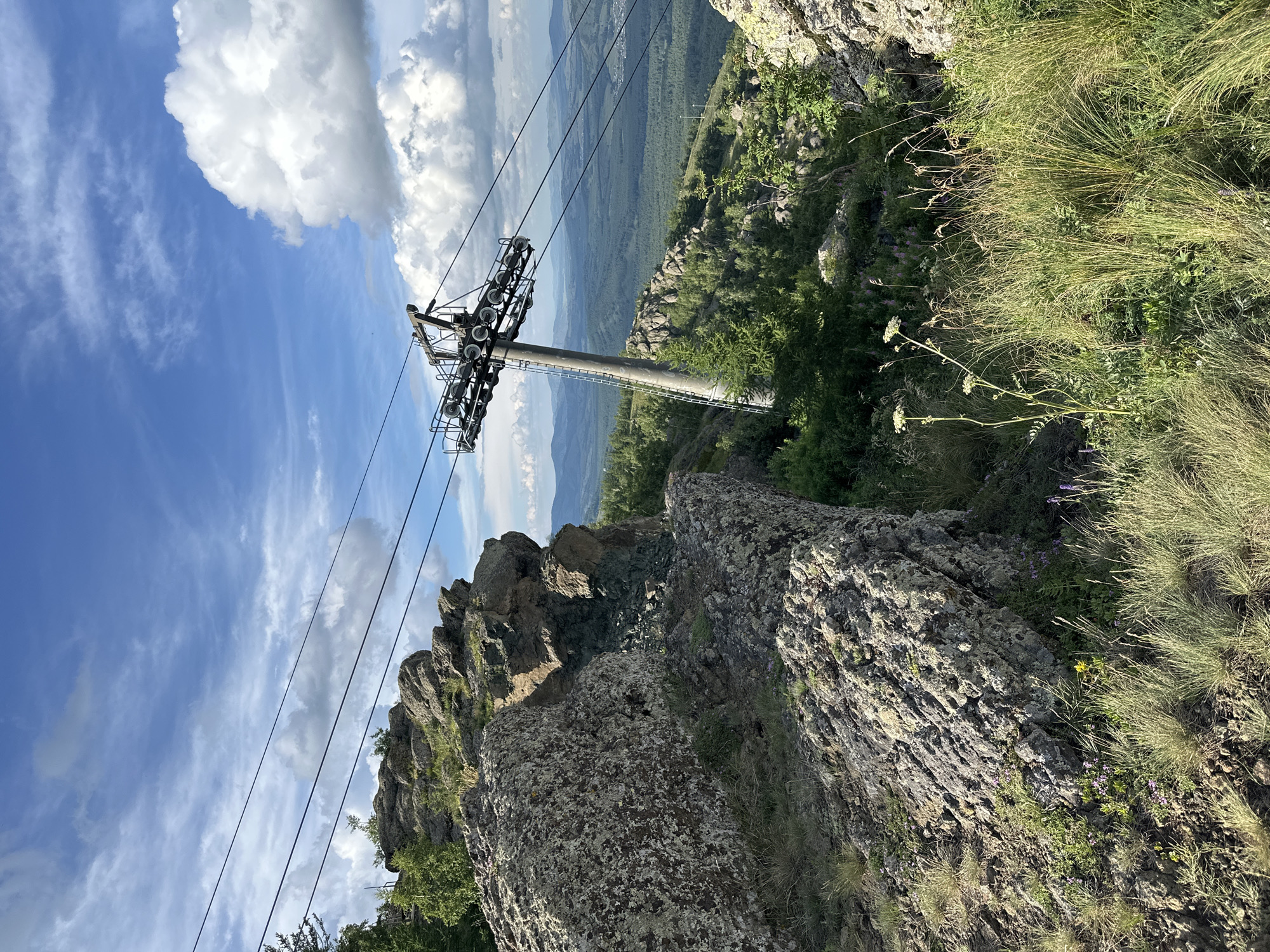 This is a medium shot, eye-level, natural light, outdoor photograph of a ski lift tower on a rocky mountain peak. The ski lift tower is a concrete structure with a metal top assembly holding multiple wheels. It is located on the right side of the frame, leaning slightly to the right. Several cables extend from the top of the tower and stretch across the frame towards the left. In the foreground, on the left side of the image, are large, rough, grey rocks covered in lichen. Further to the right and in the background, the mountain slopes are covered in green trees and smaller rocks. The sky above is blue with scattered white cumulus clouds. In the far distance, a hazy mountain range can be seen. The lower part of the image includes tall, dry grasses and some small purple and yellow wildflowers. There are no people or animals visible in the image.