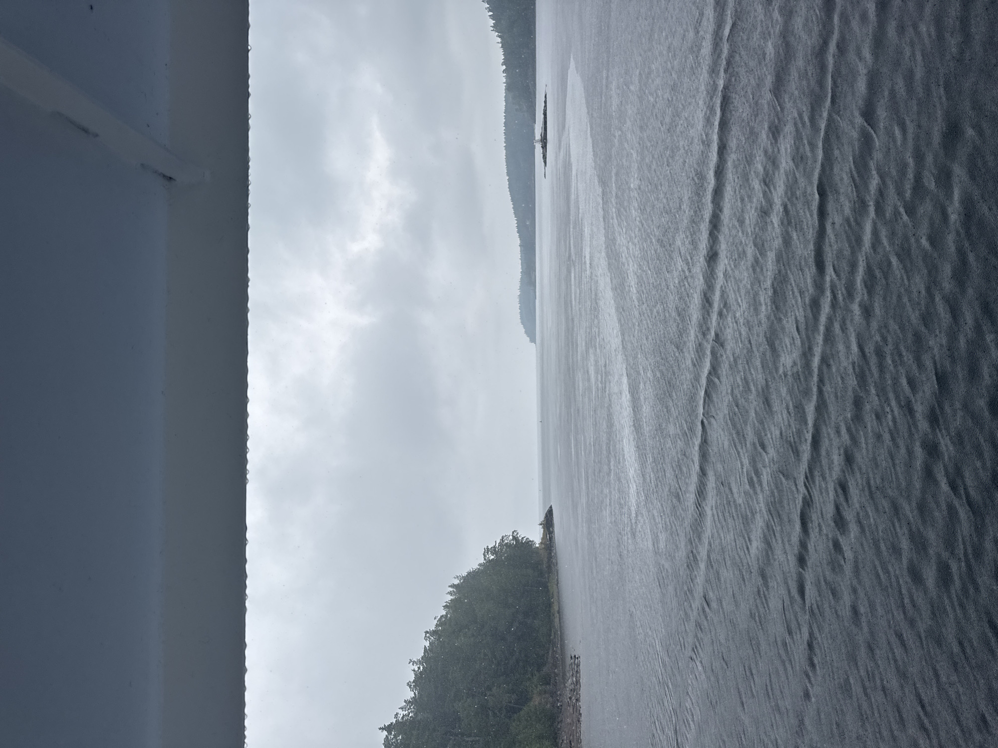 The image shows a body of water with a tree-covered shoreline on the left and a distant island with a lighthouse in the center. The sky is overcast, and it appears to be raining, with visible raindrops on the water's surface and in the air. The foreground is dominated by the dark, rippling water, with a white structure extending from the top of the frame. The location is identified as Valaam, Russia.