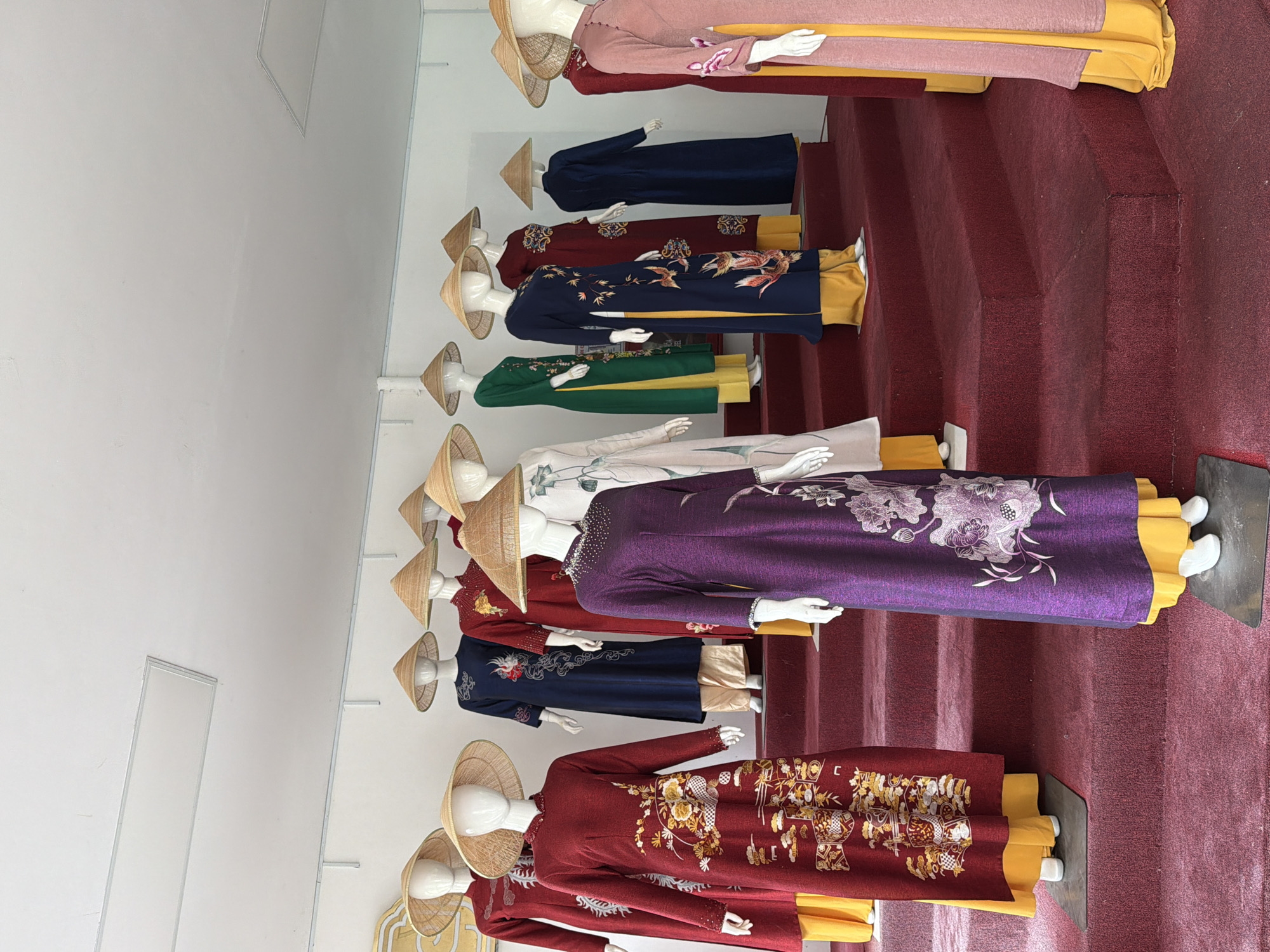 The image depicts a display of traditional Vietnamese Áo Dài dresses on mannequins, arranged on a tiered red carpeted platform within an indoor setting. The mannequins are wearing conical hats, known as nón lá, and the Áo Dài are in various colors and patterns, including deep red with gold floral embroidery, navy blue with bird and floral motifs, green, purple with floral designs, and a light pink. The background is a plain white wall with some ceiling lighting visible. The scene is likely set in a shop or museum showcasing Vietnamese culture, possibly in Thuận Hóa Ward, Vietnam, given the context. There are no people actively present in the image, only the static mannequins.