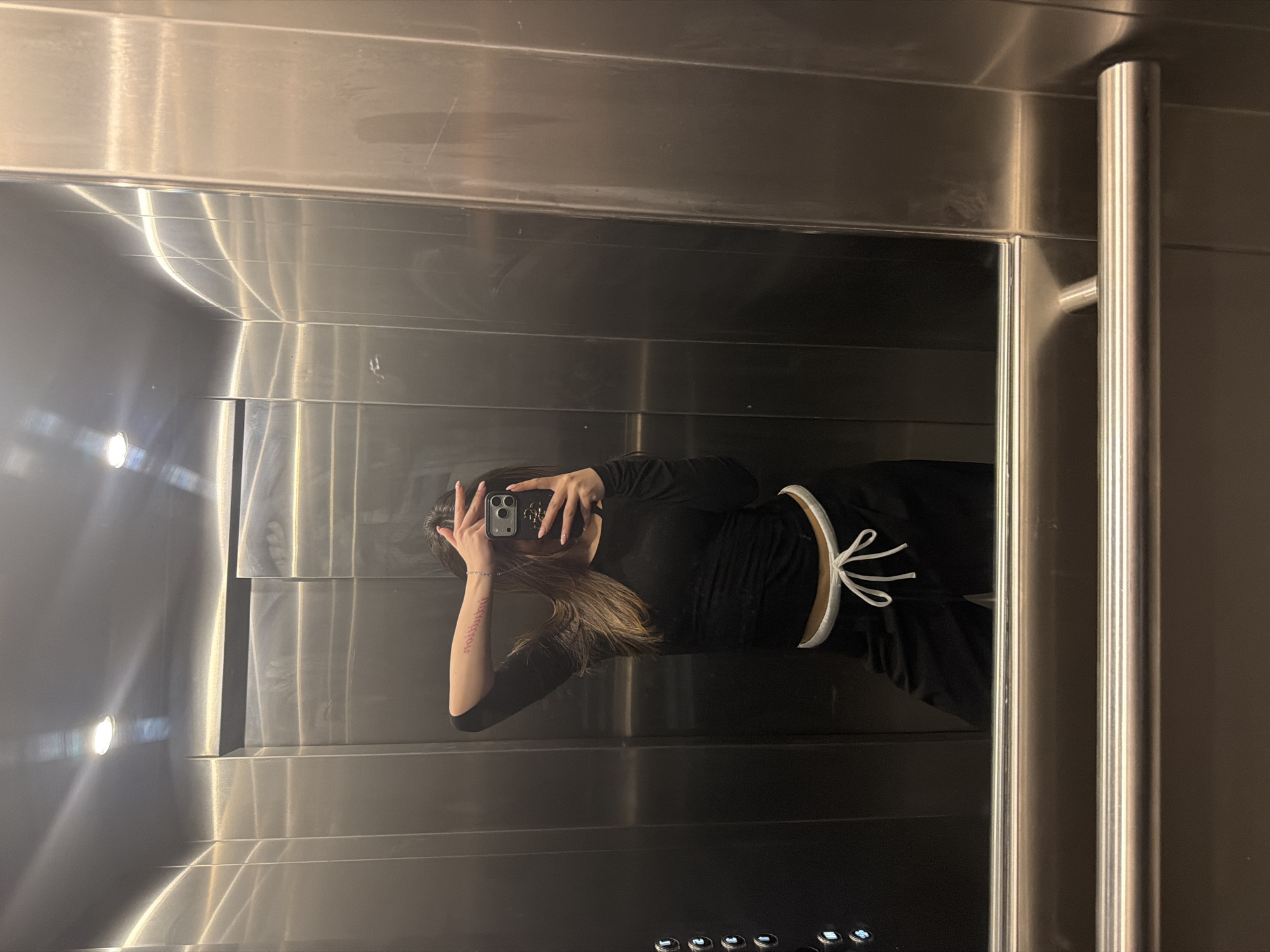 The image depicts a single individual taking a selfie inside a brushed stainless steel elevator located in Sofia, Bulgaria. The subject, a person with long brown hair, is wearing a long-sleeved black top and black pants with a white drawstring at the waist. They are holding a dark-colored smartphone in their left hand, angled towards a large rectangular mirror to capture their reflection. Their right hand is raised, partially obscuring the upper portion of their face, covering the eyes and forehead. A tattoo written in Cyrillic script, reading "София, България" (Sofia, Bulgaria), is visible on the subject's right forearm, appearing reversed in the mirror.
The elevator interior features polished brushed metal walls, with visible reflections of overhead lighting fixtures. On the left side of the reflection, a control panel with five circular, illuminated buttons is visible. A tubular metal handrail runs horizontally across the lower section of the wall below the mirror. The overall lighting is bright, with glare from the ceiling lights reflecting off the metal surfaces. The subject's fingernails are painted a light, neutral color.
