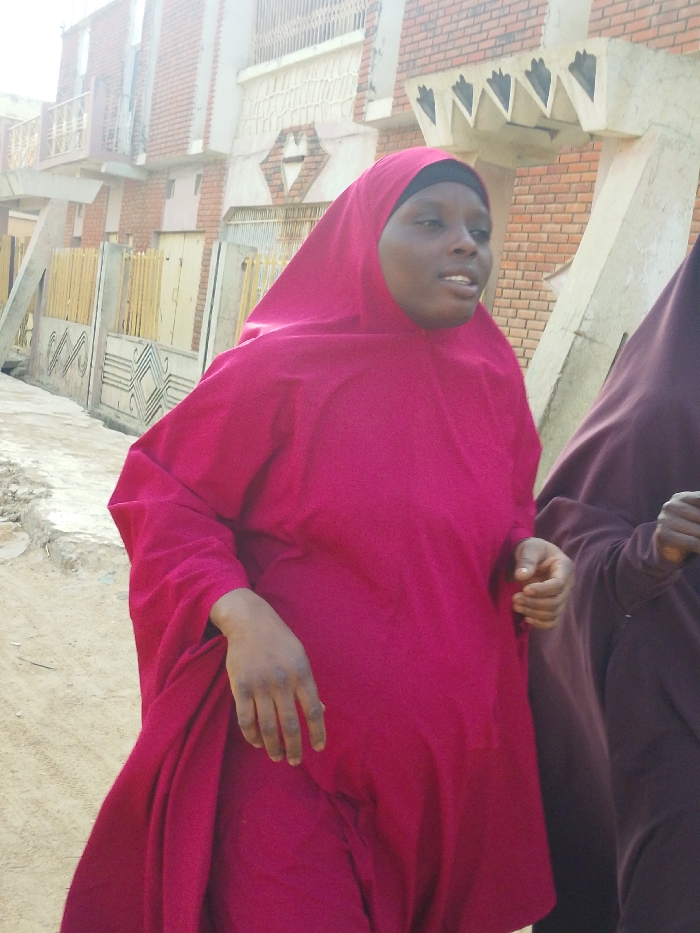 An outdoor scene in Garun Gawa, Nigeria, features a woman of color positioned centrally, wearing a bright magenta-pink hijab and a matching loose-fitting garment. She has a neutral expression with her mouth slightly open, suggesting she is speaking. Her right hand is visible, fingers naturally curled. To her right, another individual is partially visible, wearing a dark brown loose garment. The subjects are positioned on an unpaved, sandy-dirt ground. In the background, a two-story building constructed with reddish-brown bricks is visible. It features white decorative balustrades on an upper balcony, a diamond-shaped wall embellishment, and triangular concrete protrusions under the eaves. A light-colored concrete pillar stands in front of the building. A low fence or wall, possibly made of light-colored slats, and a yellowish gate or door are also discernible behind the subjects. The scene is illuminated by bright daylight.