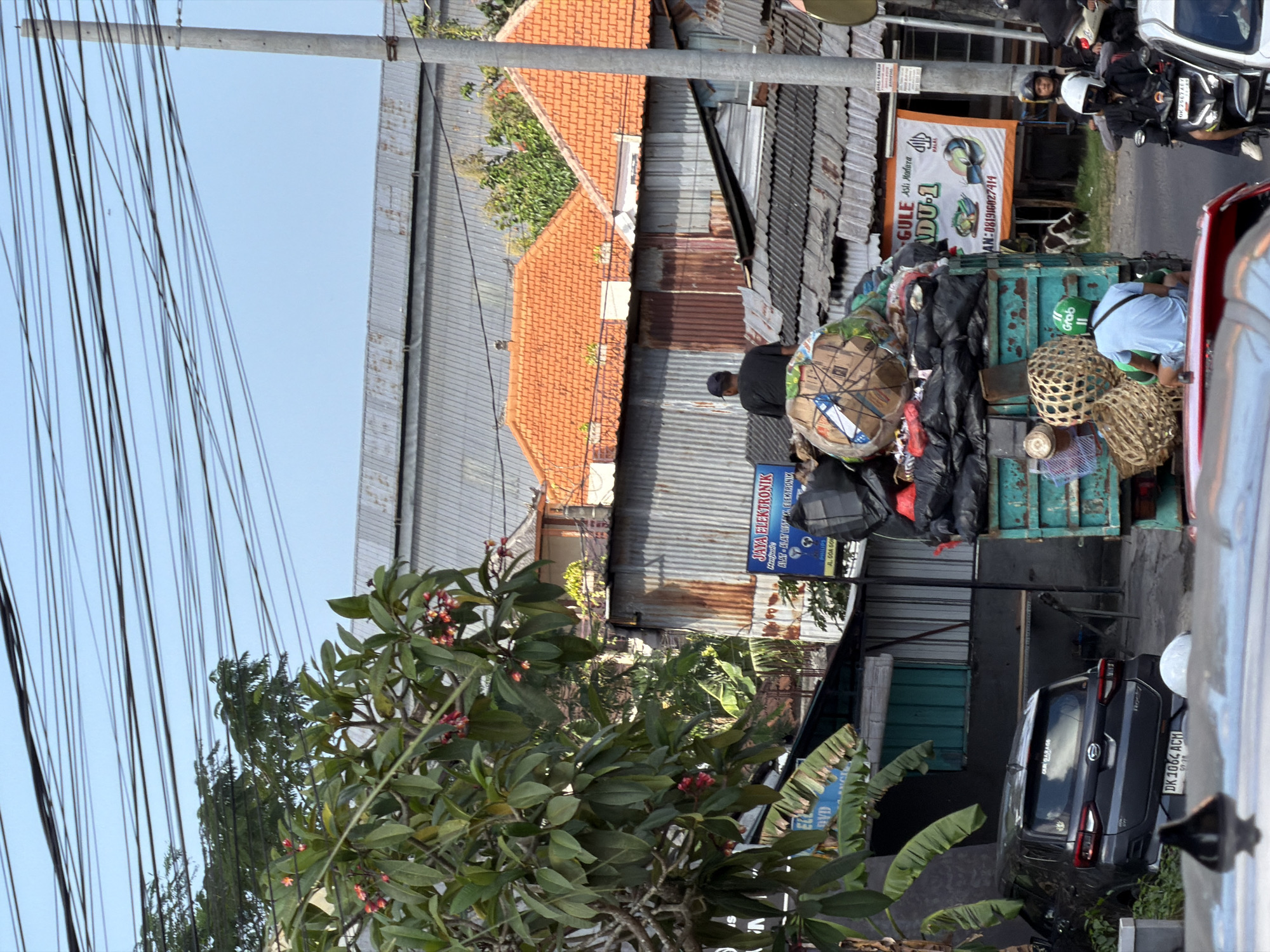 In Kuta Selatan, Indonesia, on a sunny day, a scene unfolds with a truck heavily laden with trash. The truck, positioned on a street, is a turquoise green color and its bed is overflowing with black garbage bags and other assorted refuse, including large woven baskets and what appears to be a large rolled-up tarp or carpet secured with straps. A person wearing a black t-shirt and a dark baseball cap is visible sitting atop the trash, looking towards the right side of the frame.
In front of the truck, a small dark gray SUV with license plate DK 1064 ACM is parked. A white circular object is visible behind the SUV. To the left of the SUV, a tree with green leaves and pink and red flowers provides a natural element to the urban landscape.
Further down the street, to the right of the truck, a white banner with orange and green text is displayed, featuring the words "GULE ASLI MADURA" and a logo with Arabic script. A utility pole with many wires running across it stands prominently on the right side of the image. Several motorcycles are also present in the scene, with riders wearing helmets. One motorcycle rider, wearing a white helmet and a black jacket, is stopped in traffic. Another rider on a motorcycle, wearing a green helmet, is positioned to the left of the white-helmeted rider. A cow or goat is seen grazing in the background to the right of the truck.
The background reveals a collection of buildings with corrugated metal roofs and orange tiled roofs, suggesting a densely populated area. The overall impression is a busy street scene with the prominent presence of waste management or collection.