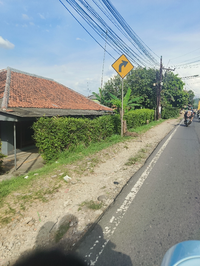 The image shows a residential street in Jatiluhur, Indonesia (latitude: -6.571, longitude: 107.44). A single-lane asphalt road with a white dividing line runs diagonally across the image, from the right foreground to the left background. A yellow road sign with a black arrow indicating a right turn is mounted on a metal pole on the right side of the road, at the edge of a narrow gravel shoulder. A residential house with a tiled roof is visible on the left side of the road, partially obscured by a green hedge. Lush vegetation grows along the roadside, and the ground is partially covered with loose gravel. A motorcycle with a rider is visible in the distance, traveling away from the camera. The scene is lit by bright sunlight and a clear blue sky. The road is empty except for the lone motorcycle. There are multiple power lines running across the scene.