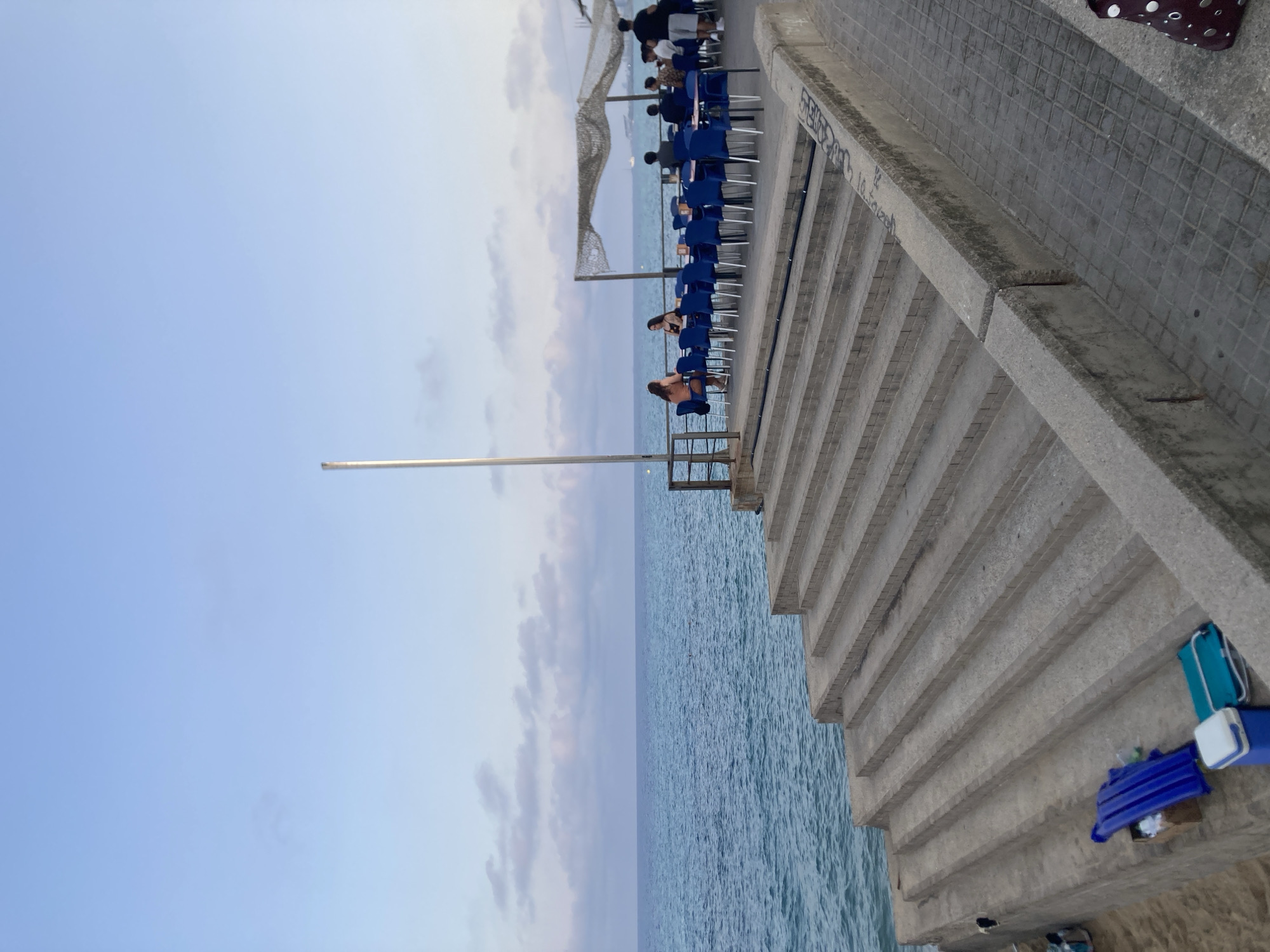 The image captures a waterfront scene at what appears to be a restaurant or cafe located in Badalona, Spain. In the foreground, a wide set of concrete stairs descends towards the sand. To the left of the stairs, several items are placed on the sand, including a blue inflatable mattress, a cardboard box, a cooler, and a folded teal bag.
Moving up the stairs and to the right, a paved walkway runs alongside a low concrete wall. Beyond the wall, a restaurant area is set up with numerous blue chairs around tables. Several people are seated at these tables, engaged in conversation or dining. The restaurant is partially covered by a canopy made of netting.
Further into the background, the turquoise Mediterranean Sea stretches to the horizon, with a hazy sky above. The water appears calm with small ripples. A tall, slender pole stands near the edge of the restaurant's seating area, and a railing separates the restaurant from the sea. The overall atmosphere suggests a relaxed evening setting by the sea.