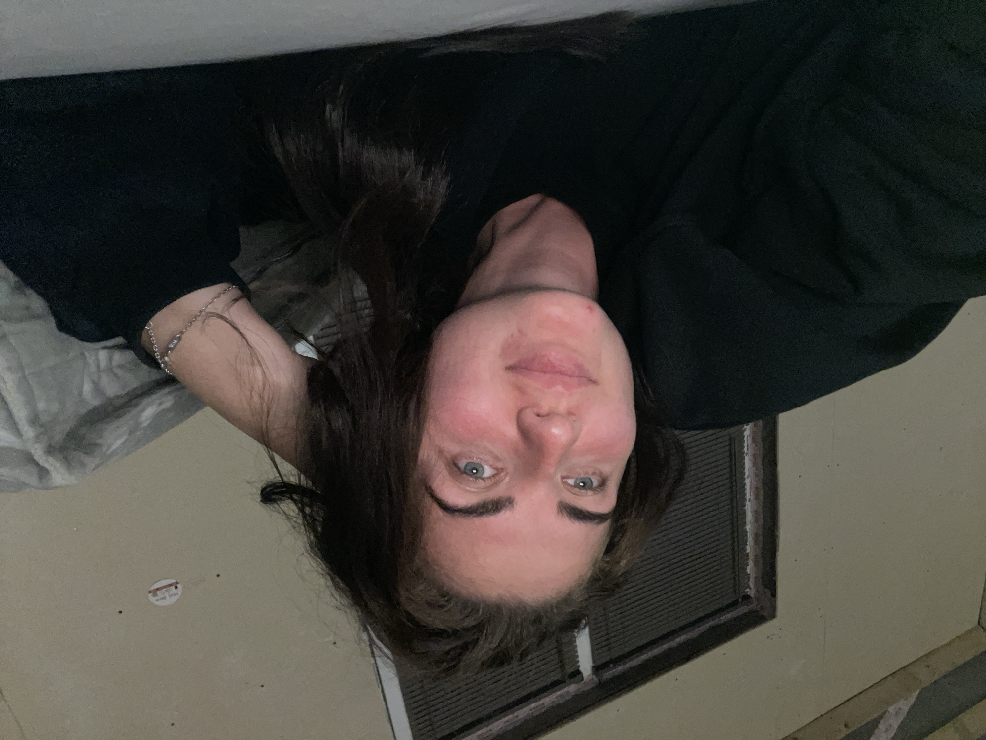 A woman is lying on a bed with her head resting on her arm, looking at the camera. She has long, dark brown hair and is wearing a black sweatshirt. Her skin appears flushed, and she has a small blemish on her chin. In the background, a window with blinds is visible, and a piece of paper is stuck to the wall above it. The room appears to be dimly lit. The scene is likely indoors, possibly in a residential setting.
