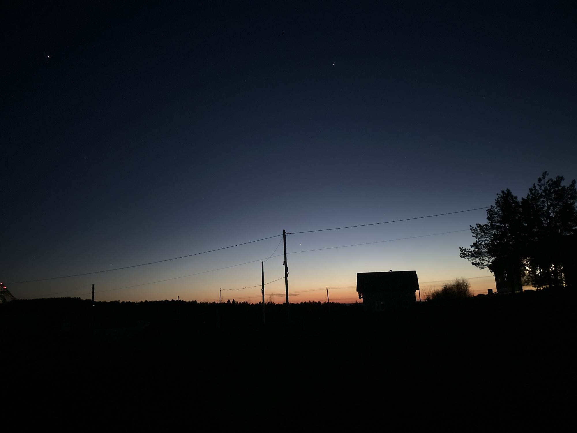 The image shows a nighttime scene in Прикамский, Russia. The sky transitions from dark blue to orange near the horizon. Silhouetted against the sky are a small building, trees, and utility poles with wires. The foreground is dark, obscuring details. Three stars are visible in the dark blue sky.