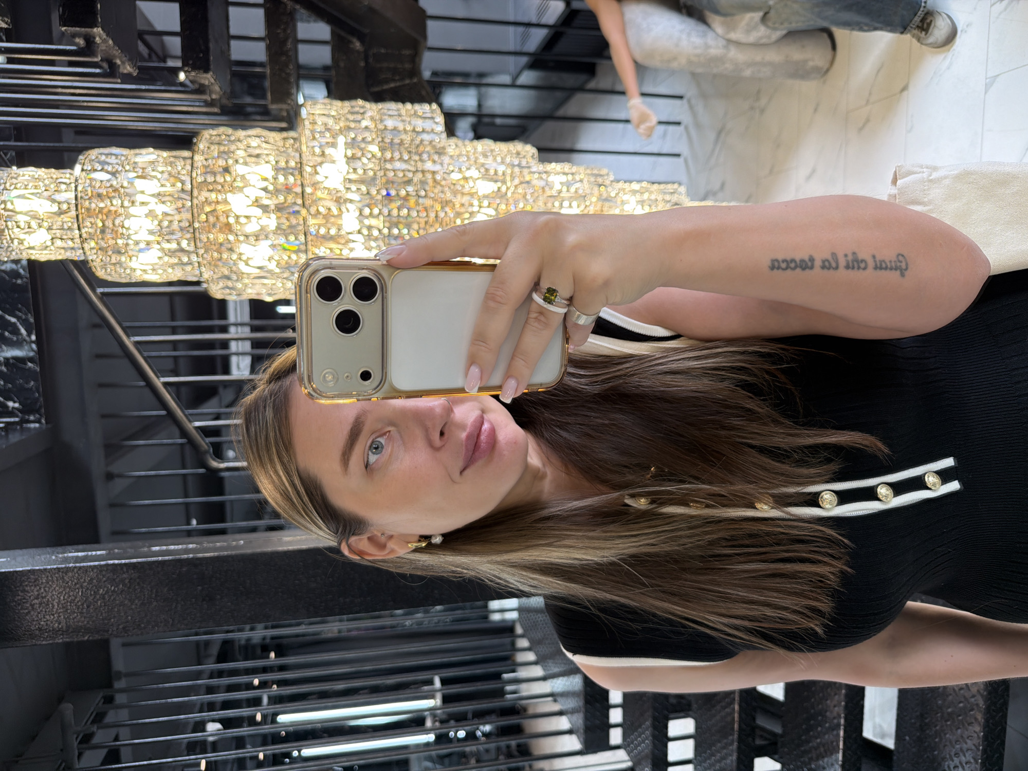 A woman is taking a selfie with her iPhone. She is wearing a black top with white and gold accents. She has brown hair with blonde highlights, blue eyes, and a tattoo on her arm. She is wearing several rings on her left hand, including one with a green stone. She is standing on a staircase with metal railings. Behind her is a large, multi-tiered crystal chandelier. The location is likely the Ritz Carlton Hotel in Moscow, Russia. In the background, a person in gloves appears to be working on the marble floor.